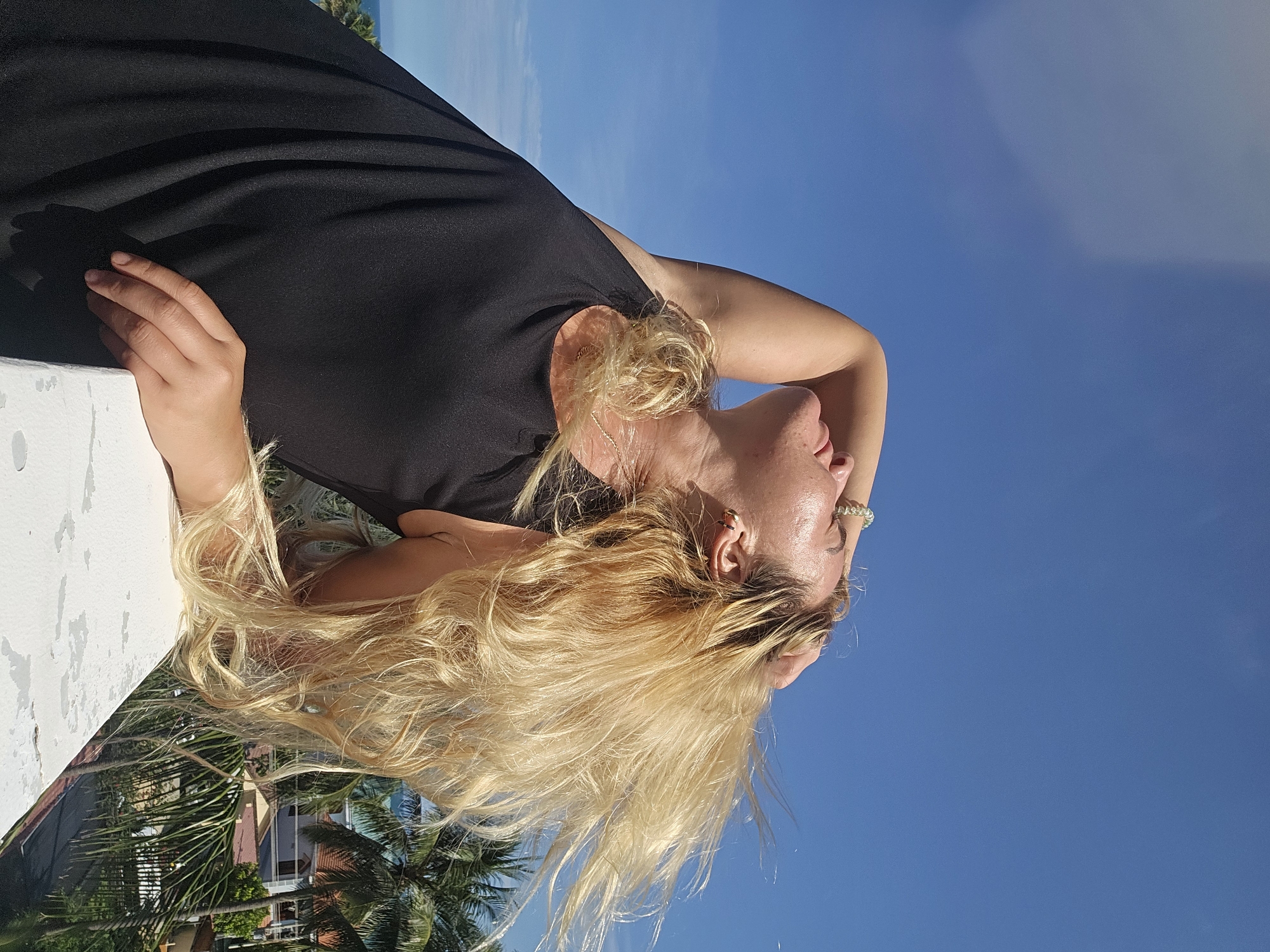 A blonde woman stands outdoors on a bright, sunny day, with her back to the camera and her head tilted back. She is wearing a black tank top and has her right arm raised, with her hand resting on her head and her hair flowing to her right. Her left arm is bent and resting on a white railing in the foreground. Behind her, palm trees and buildings are visible, suggesting a tropical or subtropical location. The sky is a clear blue with a few wispy clouds. The scene appears to be on a balcony or rooftop, offering a view of the surrounding environment.