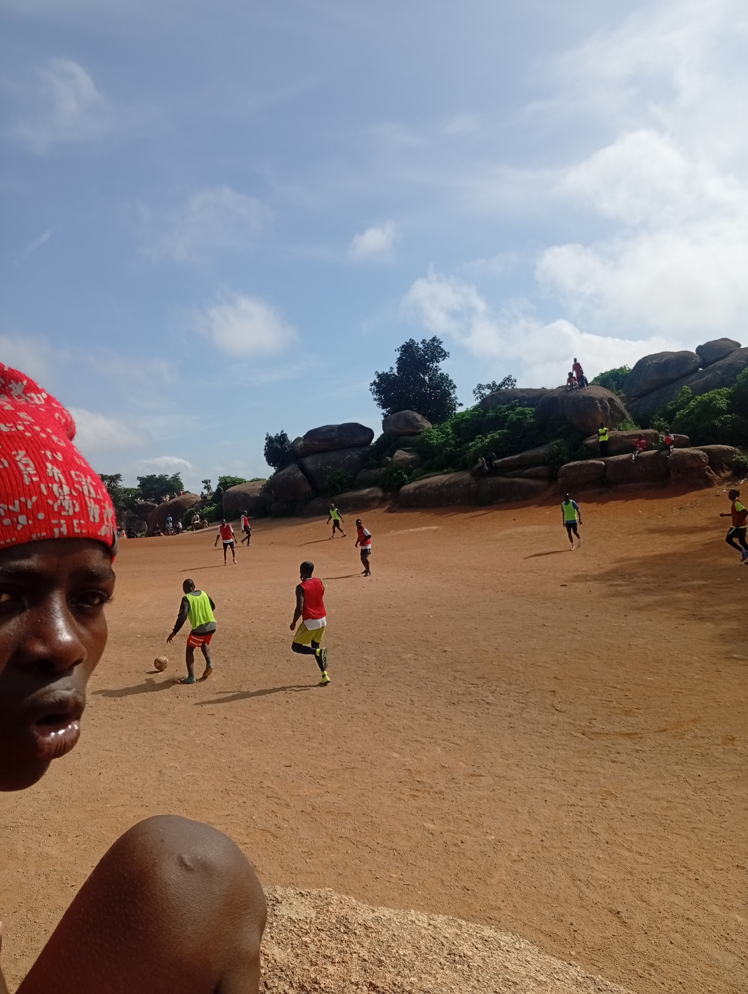 A group of young boys are playing soccer on a dirt field with large rock formations in the background. A person wearing a red beanie is in the foreground on the left, looking towards the camera. The sky is blue with scattered clouds. The scene appears to be set in a rural or semi-rural area. The overall atmosphere is one of casual recreation and community activity.