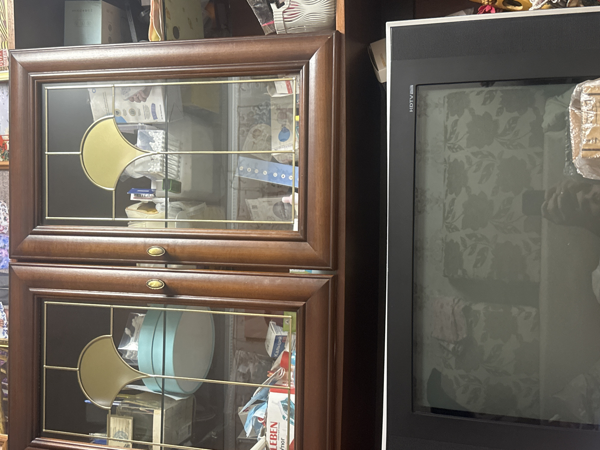 The image shows a close-up, eye-level view of a dark wooden cabinet with glass doors, filled with various items. The cabinet appears to be located in an indoor setting, possibly a living room or storage area. The items inside include boxes, bottles, and what looks like medical supplies. A pale blue, round object is prominently displayed in the left glass door. The lower portion of the image shows the top of a television screen with the "HDTV" logo visible. The background within the cabinet is dark, and some of the items are partially obscured. The scene is static and does not appear to depict any action or interaction between people. The overall impression is one of a cluttered storage space. There is no visible indication of the location being in the city of Загородный, Russia.