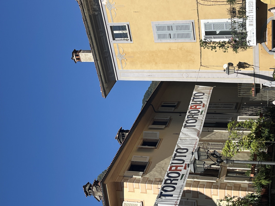This image depicts a sunlit urban scene in Varallo, Italy, specifically showing two multi-story buildings and the clear blue sky.
The building on the left is light yellow with darker yellow stone corner quoins and white-shuttered windows, some open, revealing dark interiors. It features balconies with railings and potted plants. Its roof is tiled, and a multi-tiered, ornate chimney is visible.
The building on the right is a deeper ochre-yellow with lighter yellow decorative friezes and vertical panels. It has a single window with a white frame and grilles near the top, and below that, a closed window with light blue shutters. Further down, an open window with light blue shutters reveals a balcony with railings and green climbing foliage, along with potted plants. A sign partially visible below this balcony reads "MIACCE VALSESI." This building also has a tiled roof and a simple, tall chimney.
Stretched across the street between these two buildings, a white banner with dark blue text prominently displays "TOROAUTO" repeated, with smaller text underneath indicating "VARALLO Via G. Garibaldi 39/41," confirming the precise location within Varallo.
A dark metal streetlamp with two lanterns is visible in the foreground, positioned in front of the left building, casting a shadow. Another similar lamp is partially visible on the right building. The overall scene is brightly illuminated by direct sunlight, with distinct shadows visible on the right building and the streetlamp. Distant mountains or hills are visible in the background between the buildings, under a clear, cloudless blue sky. No people are visible.