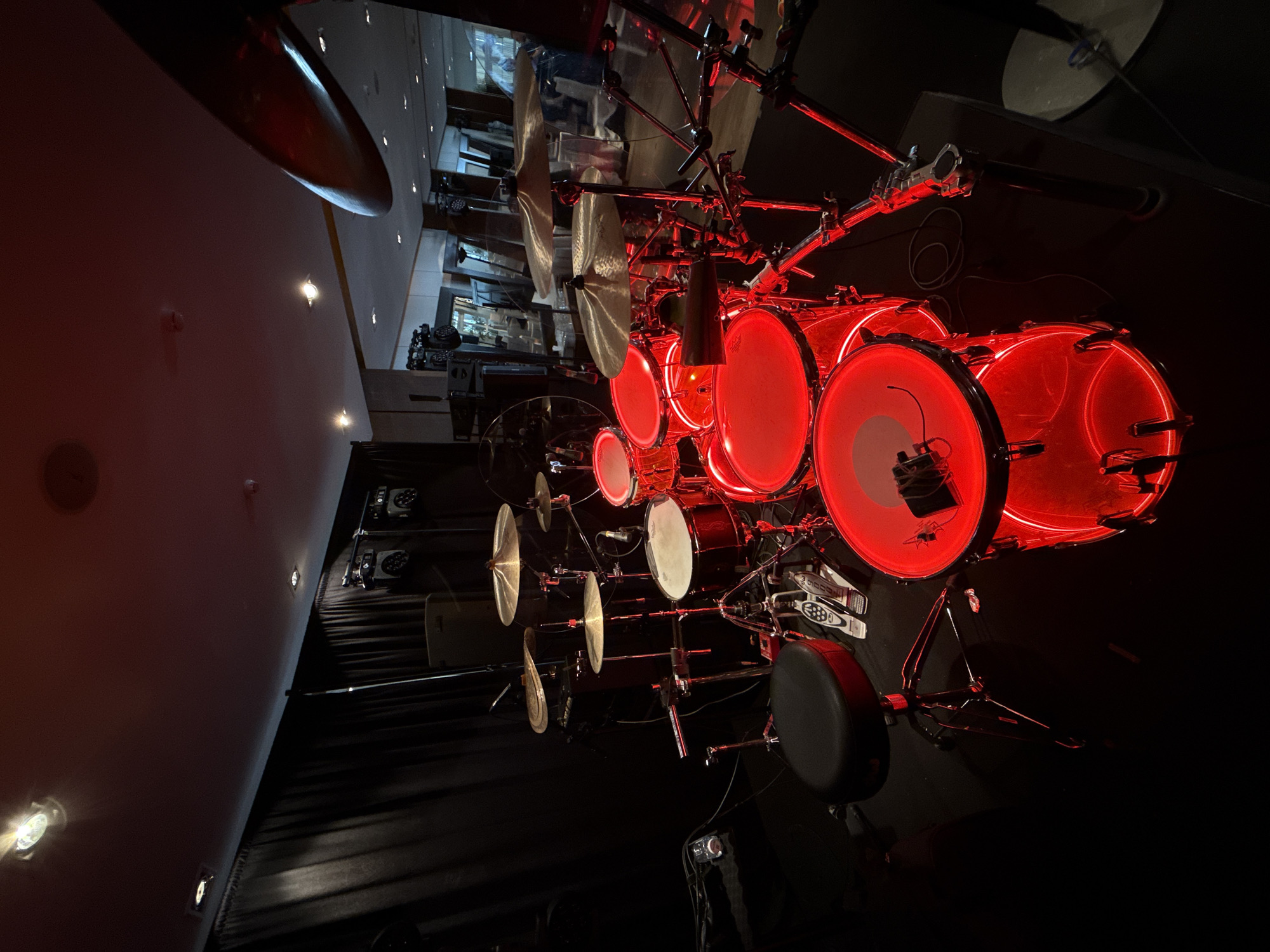 A drum set under red stage lighting is visible in a dimly lit space. The drums are red with white drumheads and there are several cymbals. A black drum stool is to the side. The drum set is in front of what appears to be a black curtain. In the background, a reflection in a glass wall shows a hallway and possibly a seating area. The environment is sparse, suggesting a stage or performance area. There is no indication of the city of Khimki, Russia.