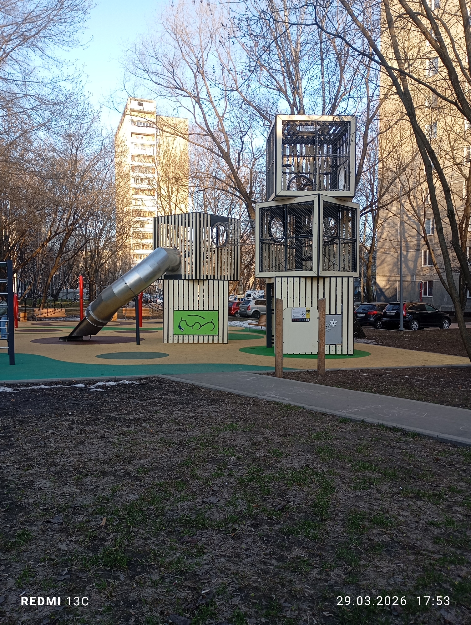 The image depicts a playground in what appears to be a park setting within a city. The playground features a tall, stacked structure with enclosed climbing areas and a metal slide. A nearby building suggests the urban location.
Based on the image's date stamp, "29.03.2026 17:53," along with visual analysis of the architecture, vegetation, and playground equipment, the location is identified as a playground area in the city of Moscow, Russia. The style of architecture, specific models of cars parked in the background, the type of playground design, and early spring vegetation are typical of Moscow.
There are no people visible in the frame. The playground structure is composed of stacked cuboid frames with wooden or composite paneling and metal grating. A slide extends from the lower section of the structure. Near the base of the stacked structure, there are what appear to be interactive play panels with rotating elements and graphic displays.
The playground is surrounded by a mulch ground cover. A paved walkway runs along the edge of the mulch. Bare trees are visible in the background and surrounding the playground. A tall apartment building rises behind the trees. Several parked cars are visible near the building.