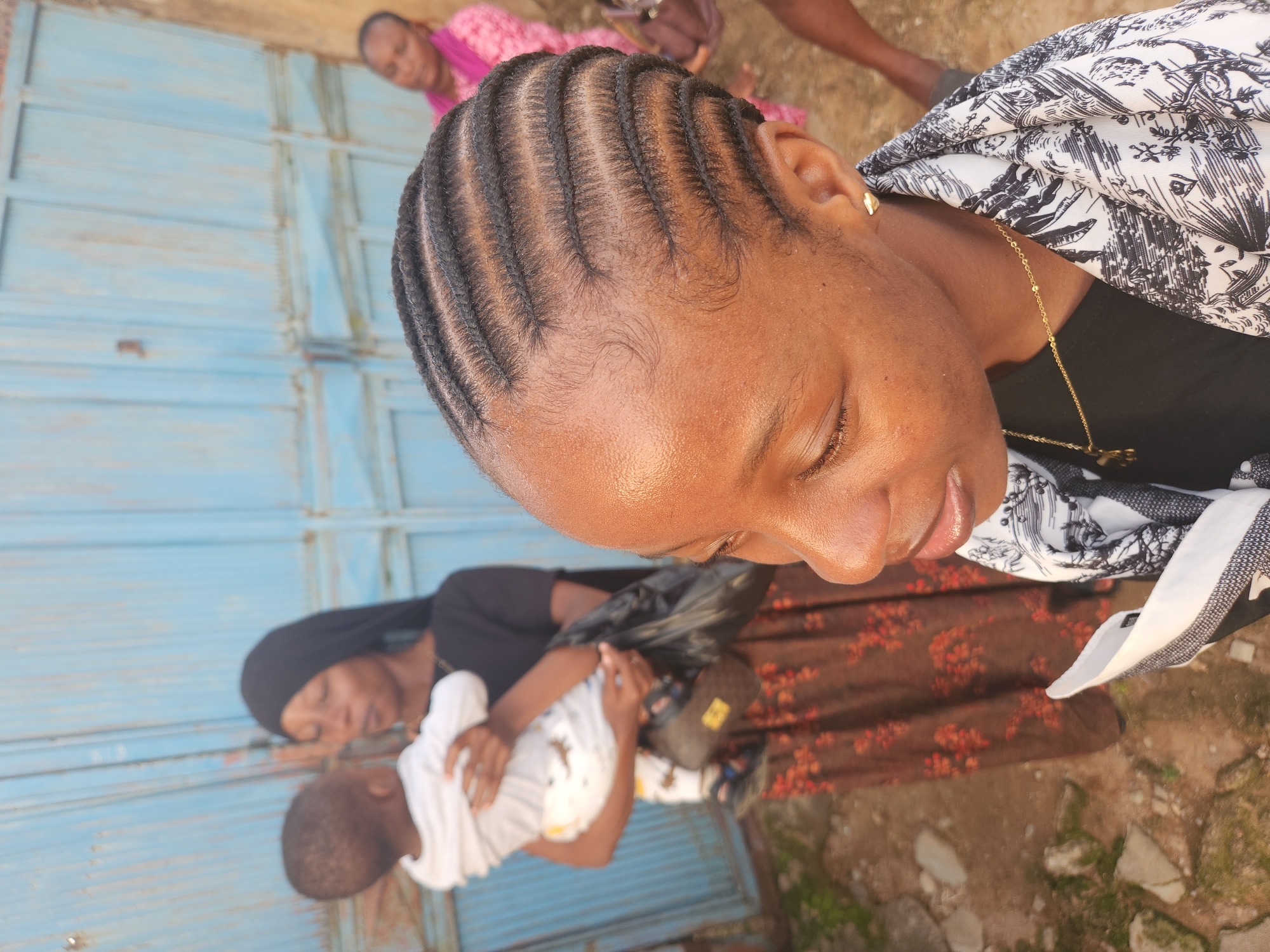 In Sabon Tasha, Nigeria, a group of people are gathered outdoors. In the foreground, a young woman with cornrows is looking down and to the right. She is wearing a black t-shirt and a scarf with a black and white pattern. A gold necklace is visible around her neck, and she has a small gold heart-shaped earring in her right ear. Behind her, a woman in a black hijab holds a baby dressed in a white outfit with a dinosaur print. The baby is facing the woman holding it. The woman in the hijab is wearing a long, floral print skirt. Another woman in a pink garment stands further in the background to the right. A blue metal gate is behind the individuals, and the ground is covered with dirt and small stones.