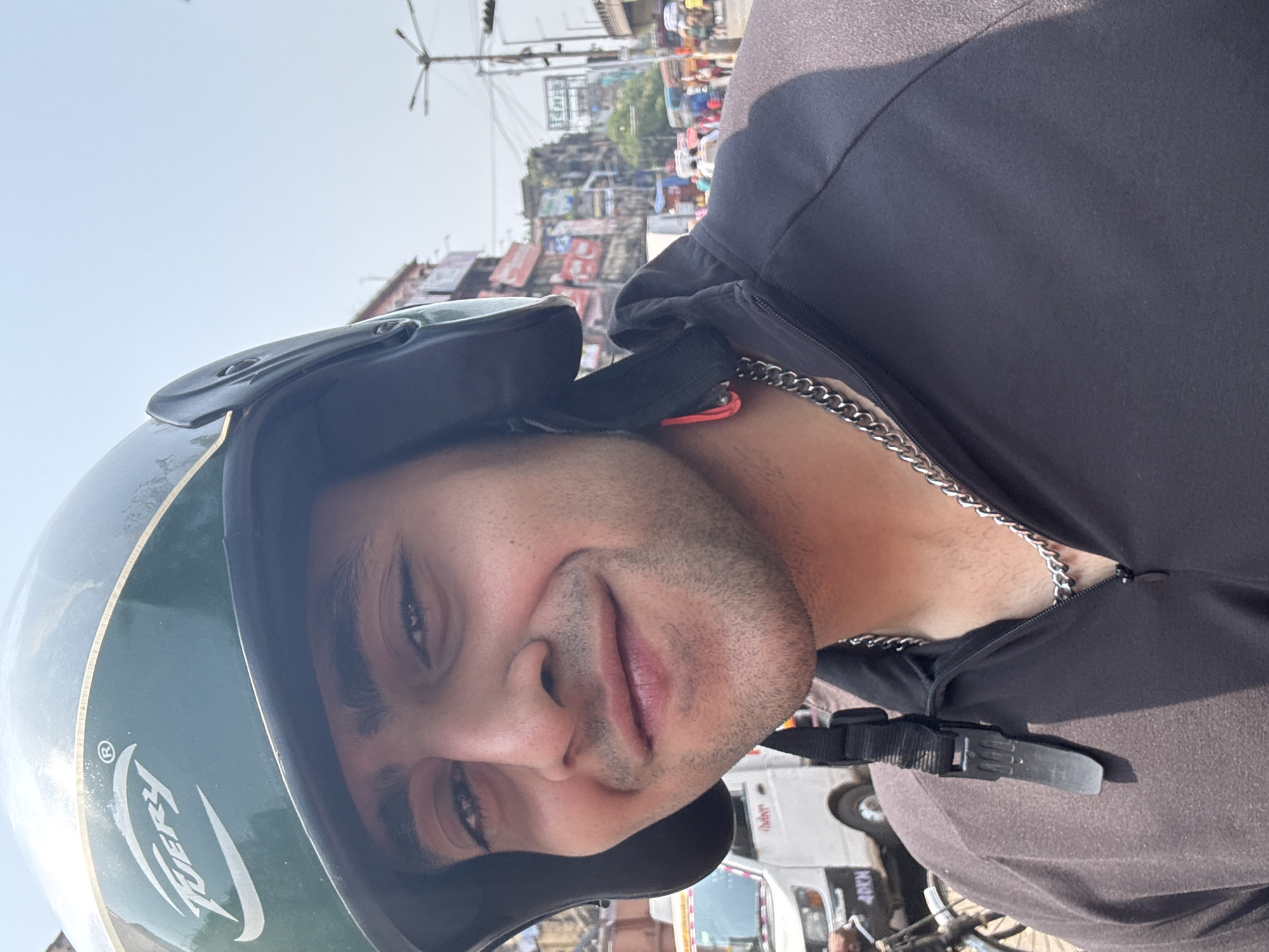 The image depicts a man wearing a green helmet and a dark-colored zip-up hoodie with a silver chain around his neck. The man is likely riding a motorcycle or scooter in a crowded urban area. The background suggests Kolkata, India, indicated by the presence of densely packed buildings, numerous street signs in an Indic script, and the general urban environment. The image captures a snapshot of everyday life in a busy Indian city.
