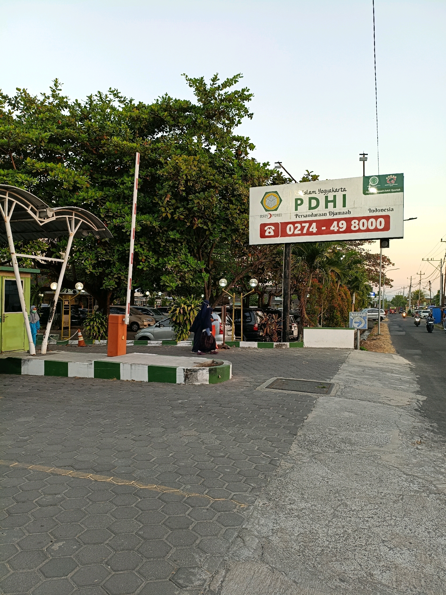 The image depicts a street scene in Purwomartani, Indonesia, with a focus on the entrance to a facility. A large white billboard displays "RS Islam Yogyakarta PDHI Persaudaraan Djamaah Indonesia" and a phone number. Two people are visible near the entrance: one in a light blue hijab and the other in a dark abaya. A red and white striped boom barrier is lowered, indicating restricted access. The ground is paved with hexagonal concrete blocks, and a grey asphalt road runs alongside. Trees and foliage are abundant, and streetlights are visible. In the background, traffic, including motorcycles, moves along the road, and power lines stretch across the sky. The overall atmosphere suggests a warm, possibly late afternoon or early evening light.