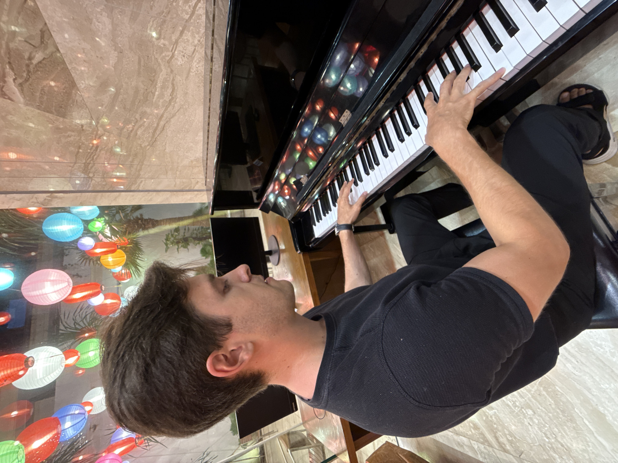 A man is sitting and playing a black piano with white keys. He is wearing a black long-sleeved shirt and black pants, along with sandals. The piano reflects a wall with decorative lanterns. The background includes marble flooring. The location is Nha Trang Ward, Vietnam.