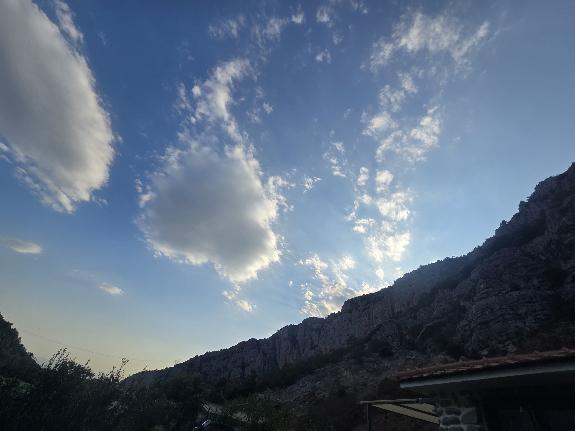 The image depicts a mountainous landscape under a blue sky with scattered clouds. A steep, rocky cliff face dominates the right side of the frame, with sparse vegetation clinging to its slopes. In the foreground, to the lower right, a portion of a building with a tiled roof and stone-clad walls is visible. Trees and dark foliage obscure the lower left portion of the image. The sky is illuminated by sunlight breaking through the clouds, creating visible rays. This scene is likely located in the outskirts of Podgorica, Montenegro, given the characteristic karst landscape. There are no people or discernible actions taking place.