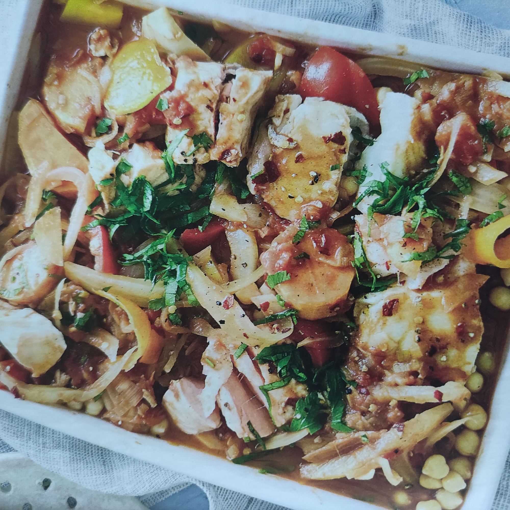 A close-up, overhead shot displays a white rectangular dish filled with a hearty stew. The stew appears to be a medley of ingredients, including chunks of white fish, sliced white onions, red tomatoes, and possibly bell peppers. The dish is garnished with chopped fresh green herbs, likely parsley or cilantro, and sprinkled with red pepper flakes. The overall color palette of the dish is a rich brown sauce with vibrant red and green accents. The dish is placed on a textured white cloth. The image is well-lit, highlighting the details of the food.