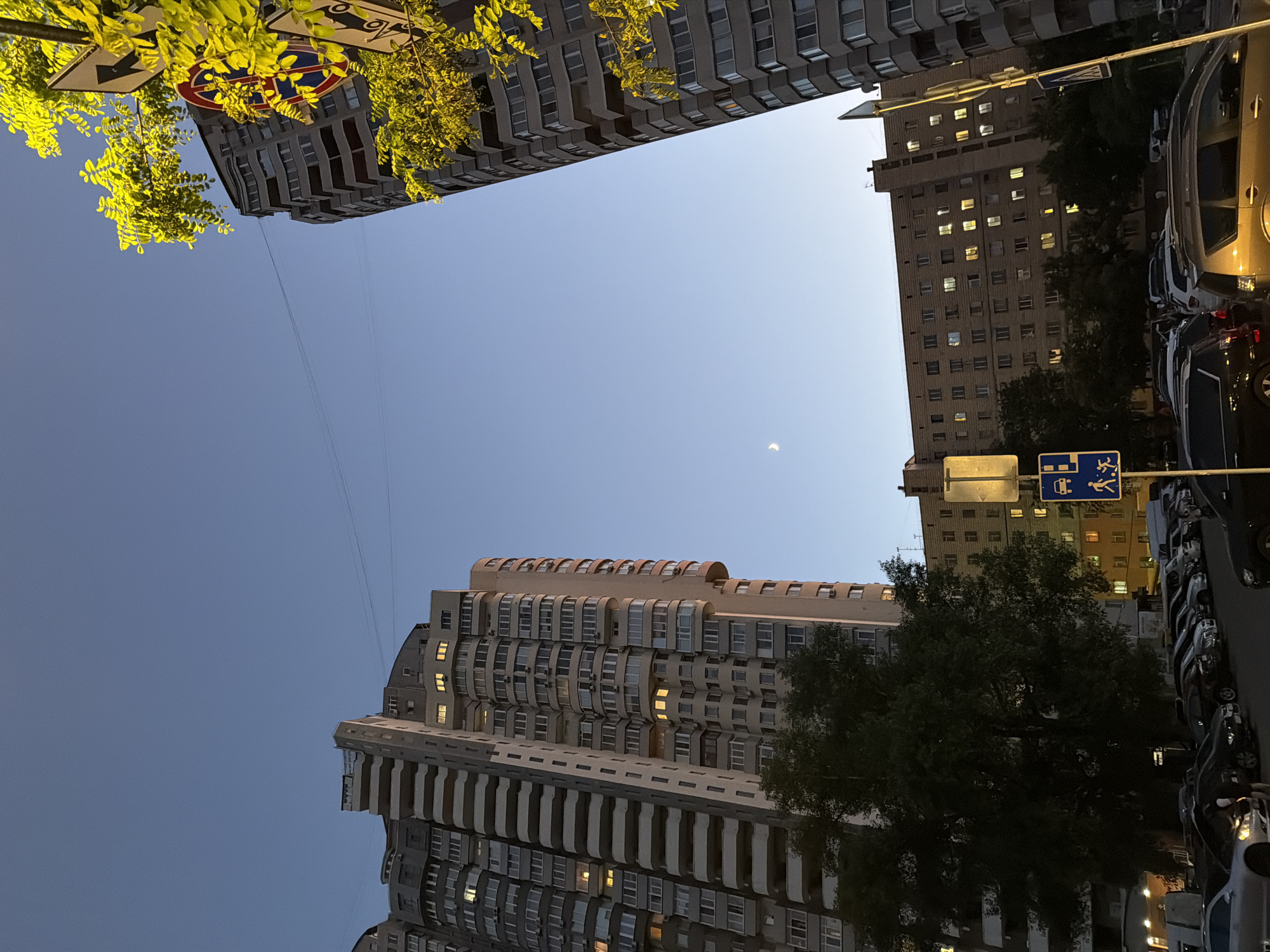 The image captures a twilight scene in Kyiv, Ukraine, dominated by residential buildings and a street with parked cars. On the left, a tall, curved apartment building with many balconies and windows is visible. To the right, another tall, grey apartment building stands, partially obscured by bright green foliage from a tree. In the background, a longer, multi-story brick building with illuminated windows stretches across the frame. The sky is a clear, deep blue, with a sliver of a crescent moon visible. The street level shows a parking lot filled with various cars, and several traffic signs are present, including one with a symbol indicating a pedestrian crossing and another with a prohibition symbol. The lighting suggests it is dusk or early evening, with some lights on inside the buildings.