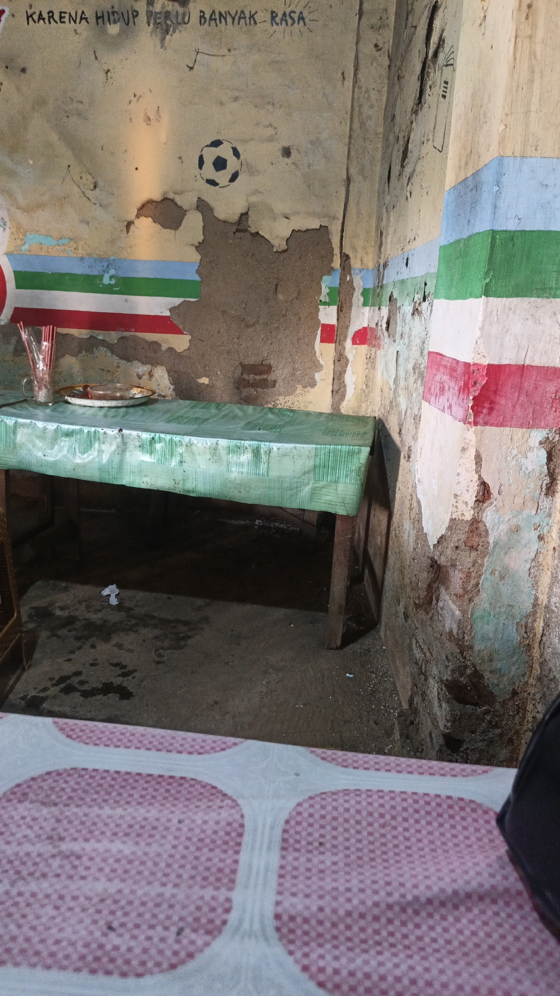 The image depicts an interior scene with peeling walls and faded paint, suggesting a potentially neglected or aged structure. In the foreground, a table is covered with a green plastic tablecloth that has a patterned texture. On the table, there is a glass with straws and a plate. The wall behind the table is decorated with horizontal stripes of red, green, and blue, and a stenciled image of a soccer ball. Above the soccer ball, there is text in what appears to be Indonesian. To the right of the soccer ball, a drawing of a simple structure, possibly a house or building, is visible on the wall. The floor appears to be concrete and is wet in places. In the lower portion of the image, a pink and white patterned surface is visible, which might be a bed or another piece of furniture. The overall environment suggests a simple, possibly humble, dwelling or establishment in the city of Dawe, Indonesia.