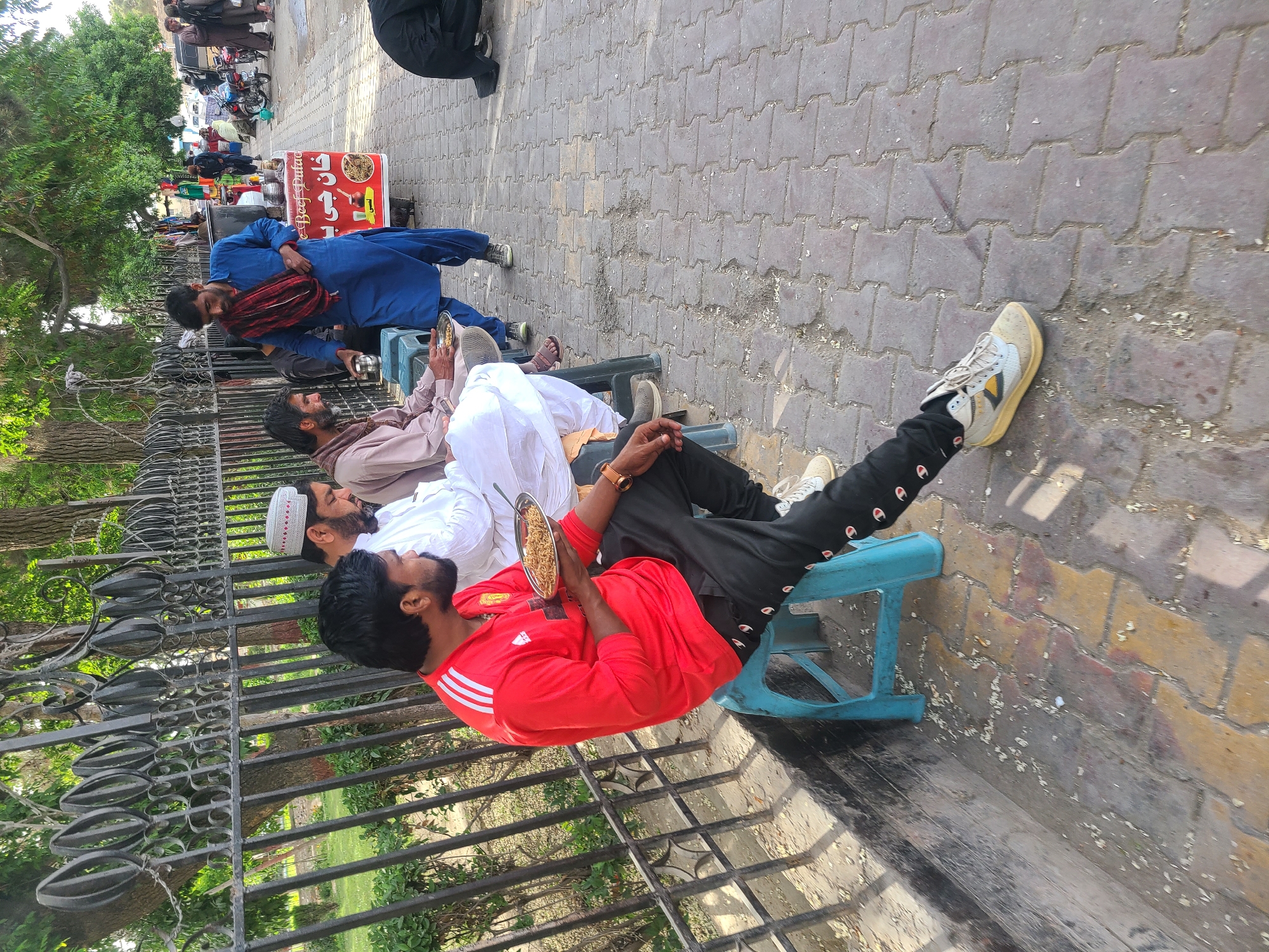 This image captures an outdoor scene, likely a busy street or public area adjacent to a park in Quetta City Tehsil, Pakistan, during daylight hours.
The main subjects are several men gathered along a black metal fence that borders a green, tree-filled park. In the foreground, four men are seated on small, light blue plastic stools. One prominent figure, wearing a red long-sleeved shirt with white stripes (possibly Adidas) and black track pants (Champion brand), is actively eating from a silver bowl, which appears to contain noodles or a rice dish. He also wears a gold-colored watch. Beside him, two other men in traditional attire (one in light brown, another in white with a white prayer cap) are seated, looking towards the right side of the frame. A fourth man, dressed in white with a prayer cap, is partially visible.
Standing slightly behind these seated individuals is a man in a blue traditional outfit (shalwar kameez) with a red and black plaid scarf draped over his shoulder. He is positioned near a red and white street food stall. This stall displays images of food and visible text in Urdu, including "گرم پکوڑے" (Garam Pakoray - Hot Fritters), "چاٹ" (Chaat), "بریانی" (Biryani), and "نورانی سموسے" (Noorani Samosay - Noorani Samosas), with "Beef Pulao" also partially visible in English.
The setting includes a paved sidewalk made of interlocking concrete blocks. In the background, more people, motorcycles, and what appear to be other small shops or vendors suggest a lively commercial or social district. The overall activity centers around eating and socializing in a relaxed, open-air environment. The time of day appears to be sunny, indicating pleasant weather.