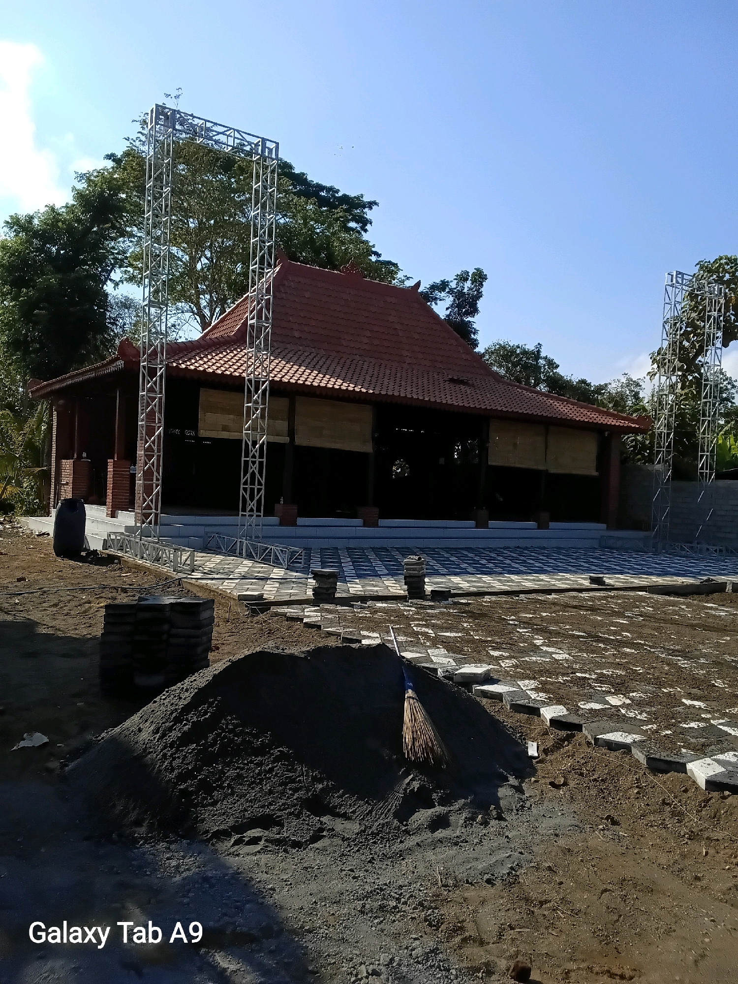 The image depicts a construction site in Trenggalek, Indonesia, featuring a traditional Indonesian-style building with a red tile roof and dark wooden accents. The building is situated in an outdoor environment with a clear blue sky and lush green trees in the background. In the foreground, there is a pile of sand with a broom resting on it, and the ground is covered with paving stones in a checkerboard pattern, indicating ongoing construction or landscaping work. Several metal truss structures, likely for event staging or lighting, are erected on either side of the building. The overall scene suggests preparations for an event or the final stages of a construction project.