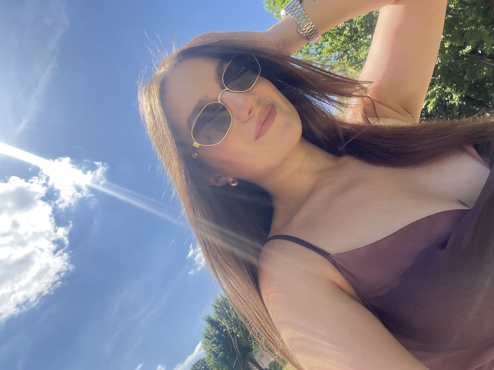 The image is a selfie taken outdoors on a sunny day with a bright blue sky and some white clouds. A woman with long brown hair is in the foreground, wearing sunglasses and a dark-colored camisole. She is resting her hand on her head, and her hair is flowing. In the background, green trees are visible. The sun is shining brightly, creating lens flares. The scene appears to be a park or natural area, possibly in the city of Ternopil, Ukraine.