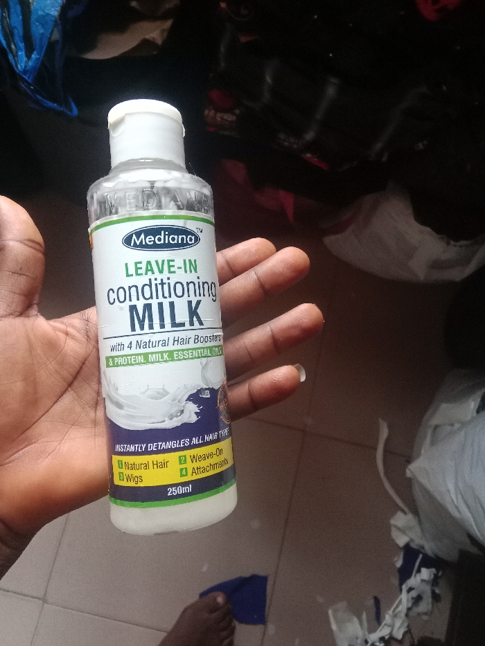 A person holds a clear plastic bottle of Mediana Leave-In Conditioning Milk. The label indicates it contains four natural hair boosters, protein, milk, and essential oils. The bottle claims to instantly detangle all hair types and is suitable for natural hair, weaves, wigs, and attachments. The product volume is 250ml. The person holding the bottle has brown skin. The background is cluttered with clothing and an indistinct floor.