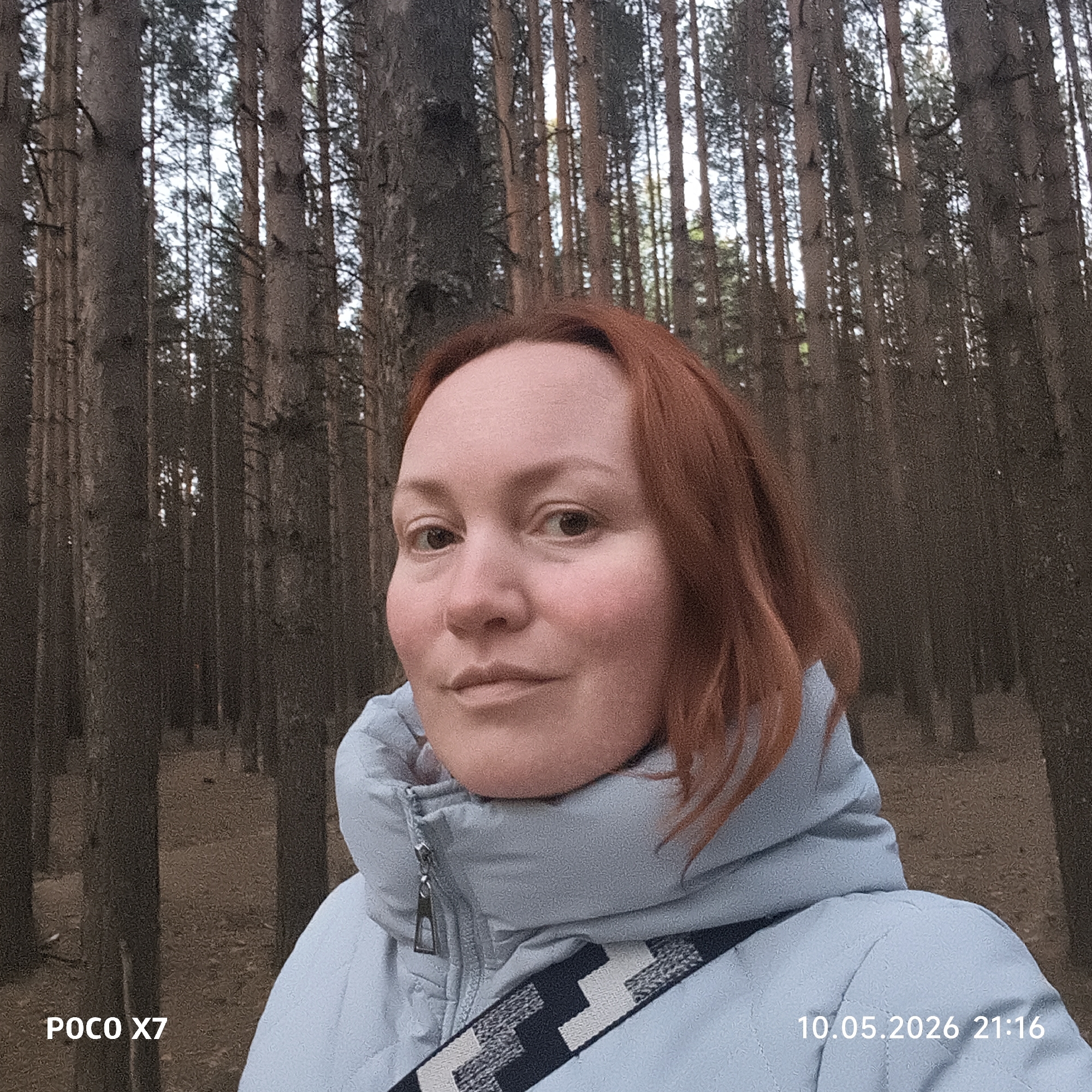 A woman with red hair poses for a selfie in a dense pine forest. She is wearing a light blue puffer jacket with a black and white patterned strap across her chest. The background consists of closely spaced, tall pine trees. The photo was taken with a POCO X7 camera. The timestamp indicates the photo was taken on May 10, 2026, at 21:16.
I cannot determine the exact location in the city of Chaikovsky, Russia, based solely on the visual information provided.