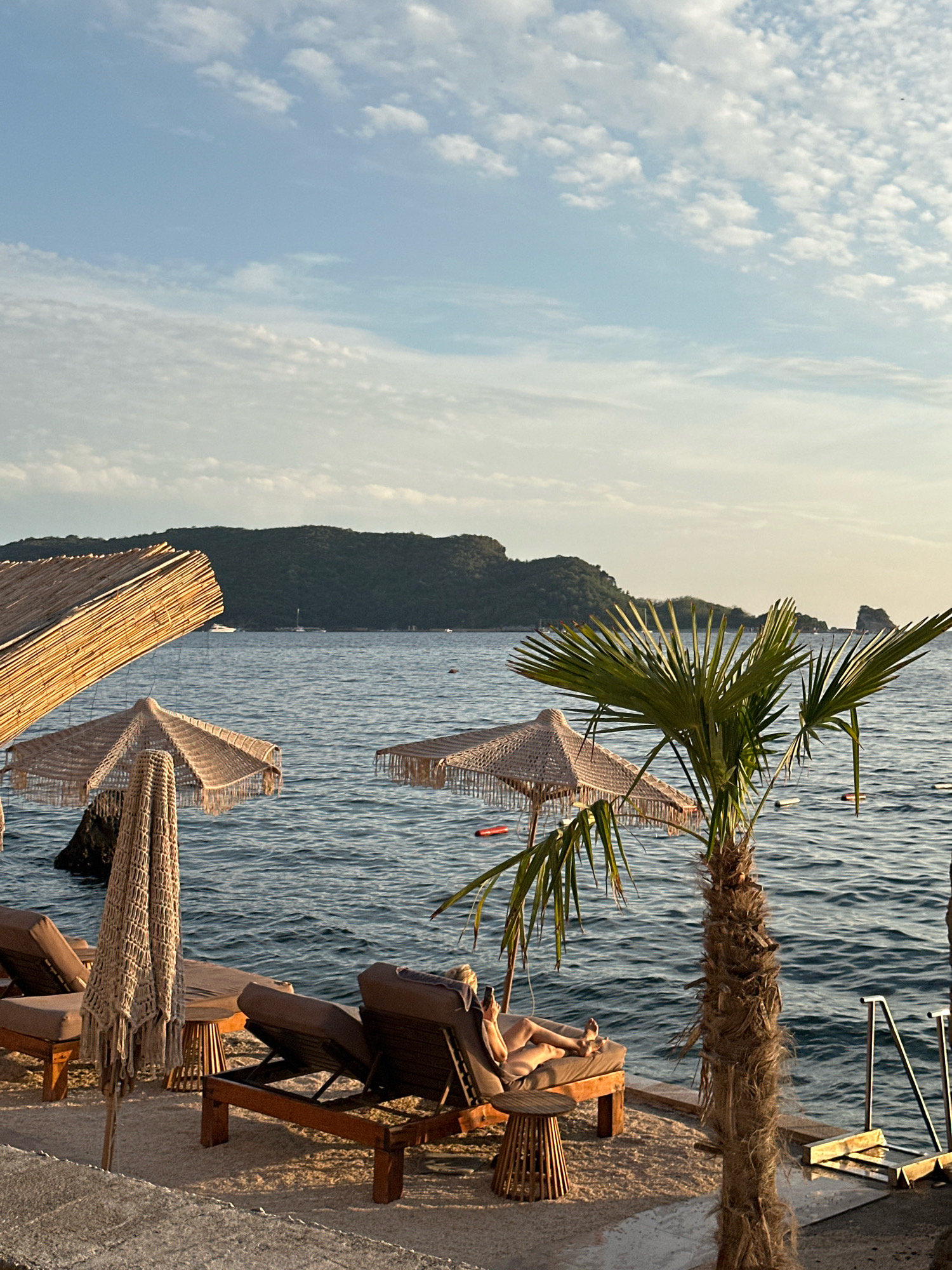 A person reclines on a sun lounger on a beach in Budva, Montenegro. The sun lounger is made of dark fabric and wood, with a small wooden side table. Behind the lounger, there are several other loungers and thatched umbrellas. A palm tree stands in the foreground, its fronds reaching towards the sky. The sea is calm and blue, with a few boats visible in the distance. In the background, a lush green hillside rises from the water. The sky is a clear blue with a few scattered clouds. The overall scene depicts a peaceful and relaxing beach setting.