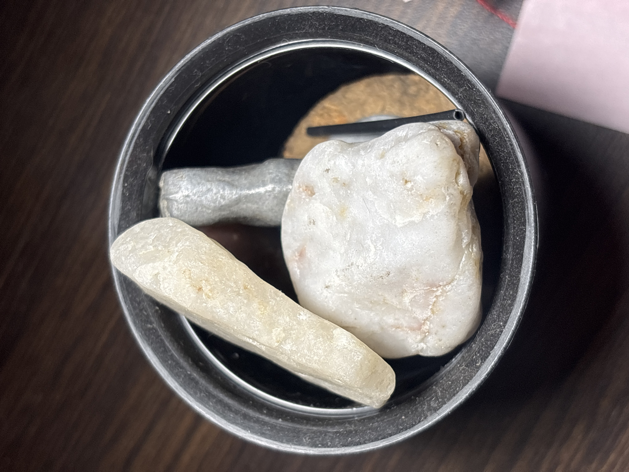 A top-down shot depicts the interior of a metal container holding three rocks. One rock is white and cone-shaped, another is a larger, rounded, off-white rock, and a third is a smaller, gray, cylindrical rock. A thin, black object sits near the center, with a tan object behind it, all within the container. The container rests on a dark, wood-grain surface. The edges of a pink object are visible in the upper right corner. The location is not discernible in the image.
