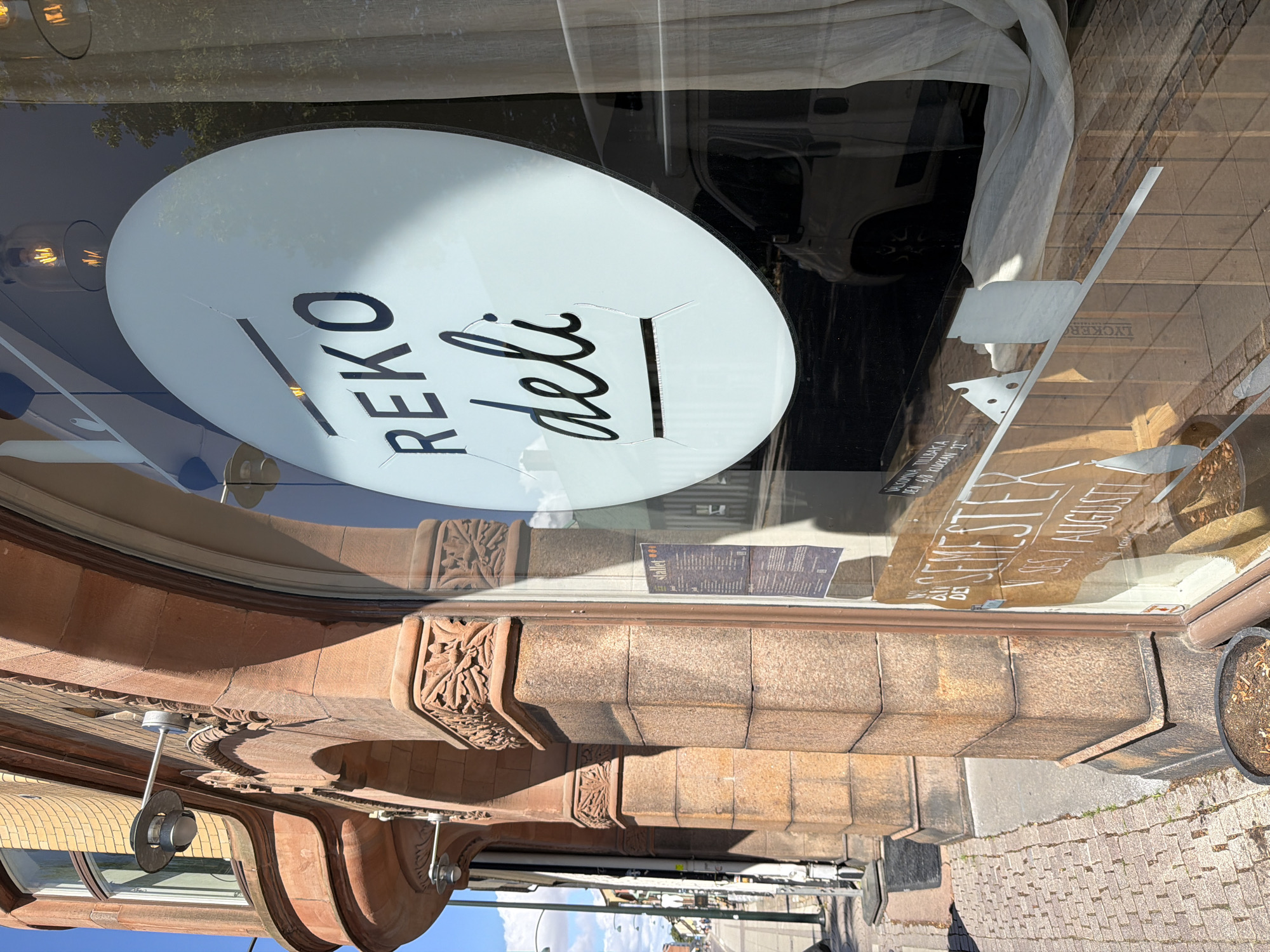 The image shows the exterior of a building in the city of Lund, Sweden. The building's facade is made of reddish-brown stone and features arched windows. On the right side of the image, a large circular sign with the text "REKO deli" is visible through the glass window of a shop. The sign appears to be made of white material and has some cracks. Inside the shop, a white curtain is partially visible. Reflections on the glass show trees and a white vehicle. A menu or flyer is attached to the inside of the window. The sidewalk in front of the building is paved with cobblestones. There are streetlights and a potted plant. The sky is blue with some clouds. The overall scene depicts a commercial storefront on a sunny day.