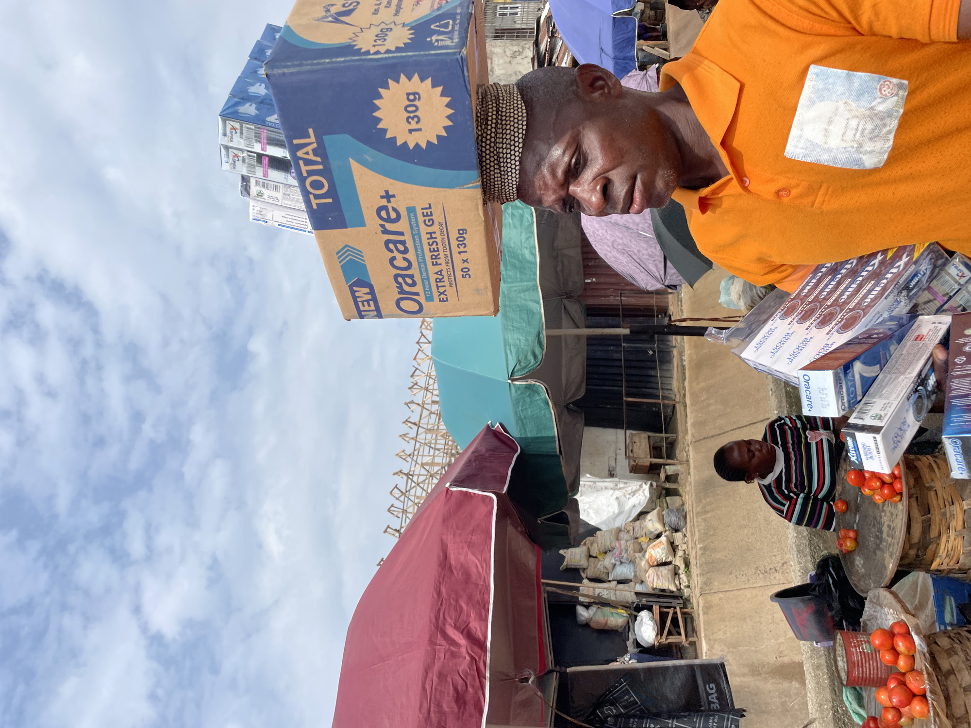 The image depicts an outdoor market scene under a cloudy sky. In the foreground, a Black man wearing an orange polo shirt balances a large box of "Oracare+" dental protection system on his head. His shirt has a visible pocket with an image of a man's face. Behind him, various market stalls are visible, some covered by large red and green umbrellas. In the middle ground, a woman with braided hair sits behind a table laden with tomatoes and other goods, wearing a striped t-shirt. Bags of goods and produce are stacked around the market stalls. The environment suggests a bustling marketplace, possibly in a developing country, with a mix of goods and people engaged in commerce.