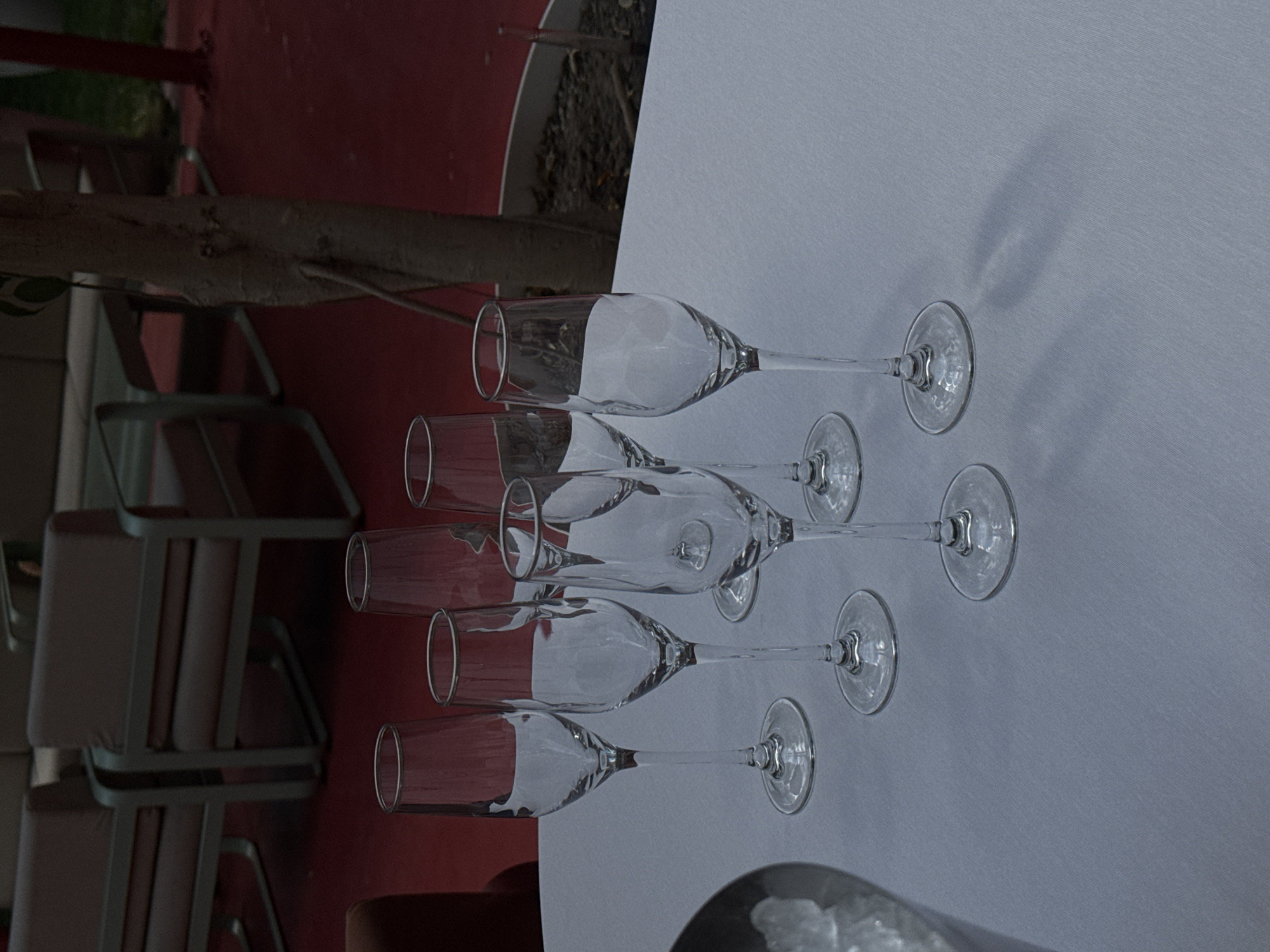 A horizontal, high-angle shot depicts five champagne flutes lying on their sides on a white tablecloth. The flutes are arranged in a staggered row, with the bowls aligned and the stems extending in parallel. Light reflecting off the glass creates highlights and shadows on the surfaces of the flutes and tablecloth.
To the left, a segment of a tree trunk is visible, followed by a series of red surfaces and the backs of several chairs with metal frames. In the lower right corner, part of an ice bucket filled with ice is visible. The context of Yerevan, Armenia, is not discernable from the given visual information.