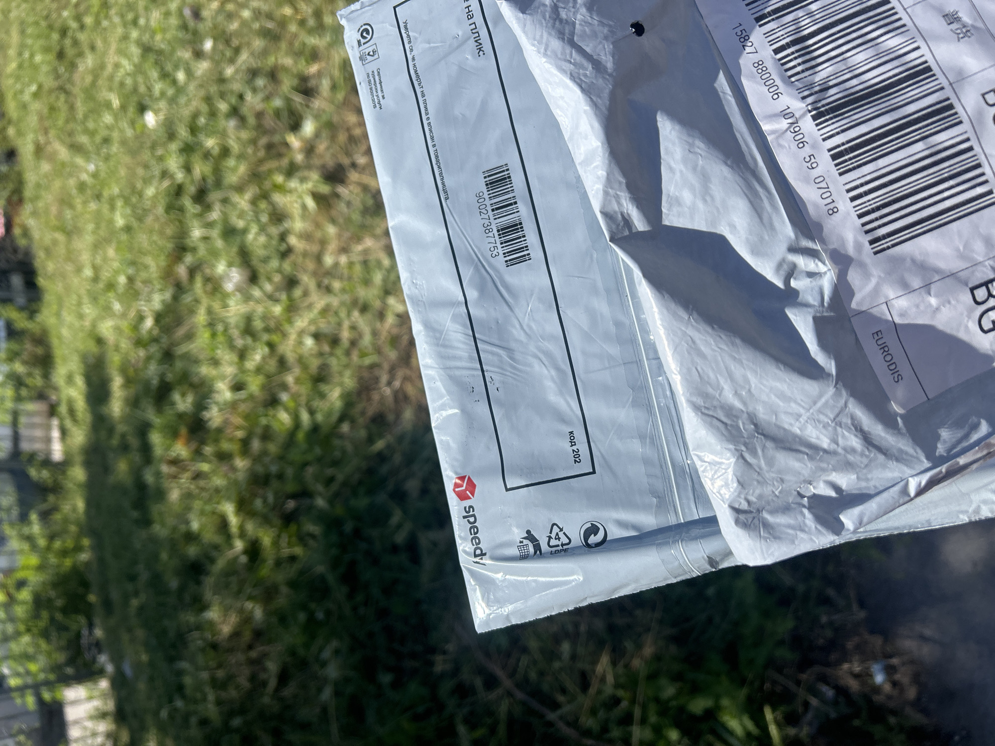 The image shows two envelopes held outdoors, with a background of green grass and foliage, suggesting a park or garden setting. One envelope, on top, is made of white plastic and has the "Speedy" logo, a Bulgarian courier company. It features a barcode and the number "90027387753". Below this, a second envelope, also white, has a barcode and the number "15827 880006 59 07018". The presence of the Speedy logo indicates the location is likely in Bulgaria, as this is their primary service area. Ruse, Bulgaria, is a plausible location given the prevalence of such courier services in urban areas.