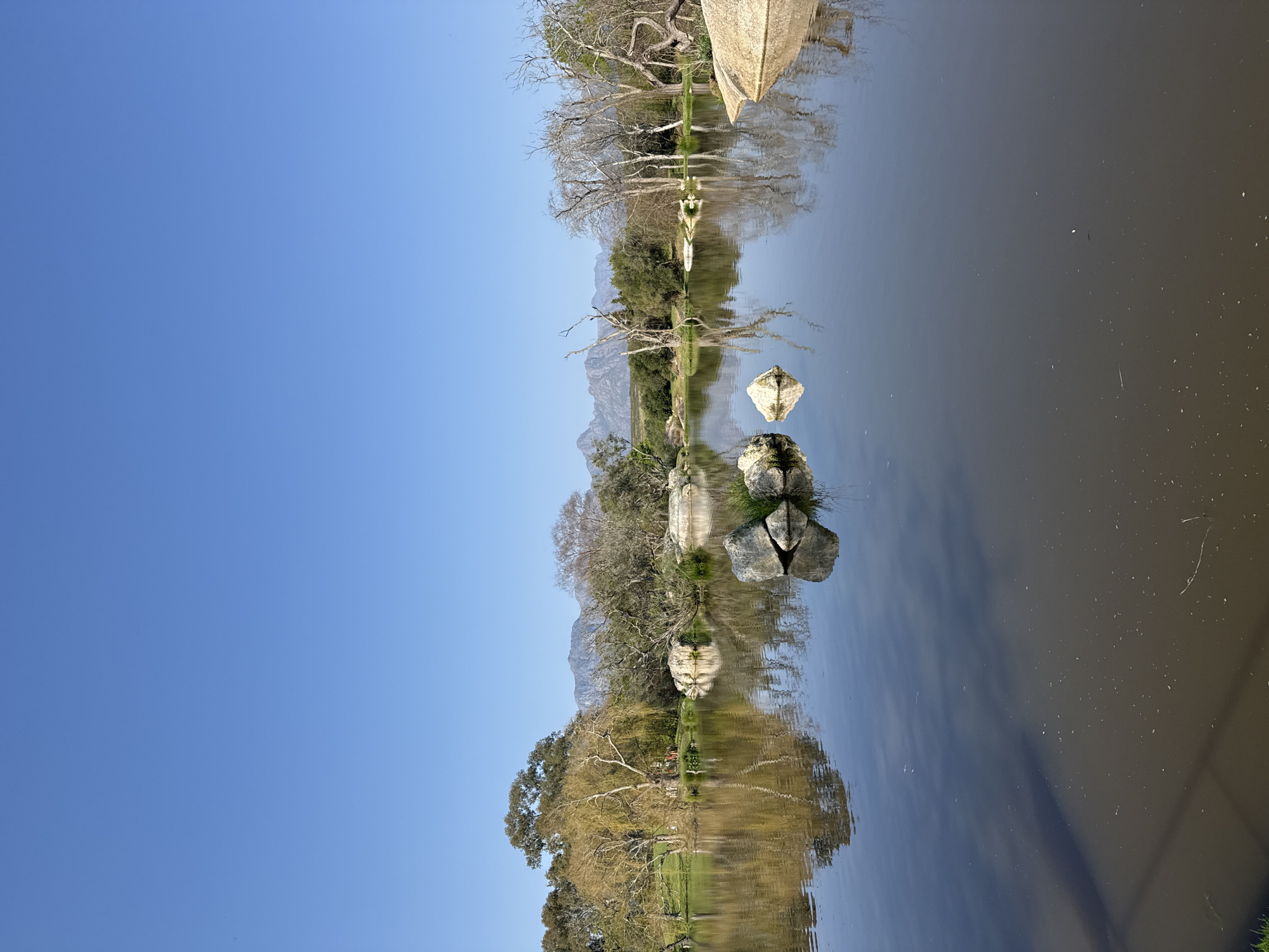 The image depicts a serene natural scene with a calm lake reflecting the surrounding landscape under a clear blue sky. The water is a dark, muted color, with visible ripples and small debris on its surface. Several large rocks are scattered in and around the lake, some emerging from the water while others rest on the shore.
The shoreline is characterized by lush green grass and a variety of trees. Some trees are bare and skeletal, likely deciduous trees in a dormant state, while others are full and leafy, suggesting a mix of species and seasons. The trees create a natural border around the lake, with their reflections mirrored in the water. In the background, a range of mountains forms a majestic backdrop, their peaks appearing somewhat hazy against the bright sky.
The overall impression is one of tranquility and natural beauty, possibly in a park or a nature reserve. The location is identified as Stellenbosch Nu, South Africa. There are no people or animals visible in the image, and the scene is static and undisturbed. The lighting suggests it is daytime, with the sun casting a gentle illumination on the landscape.