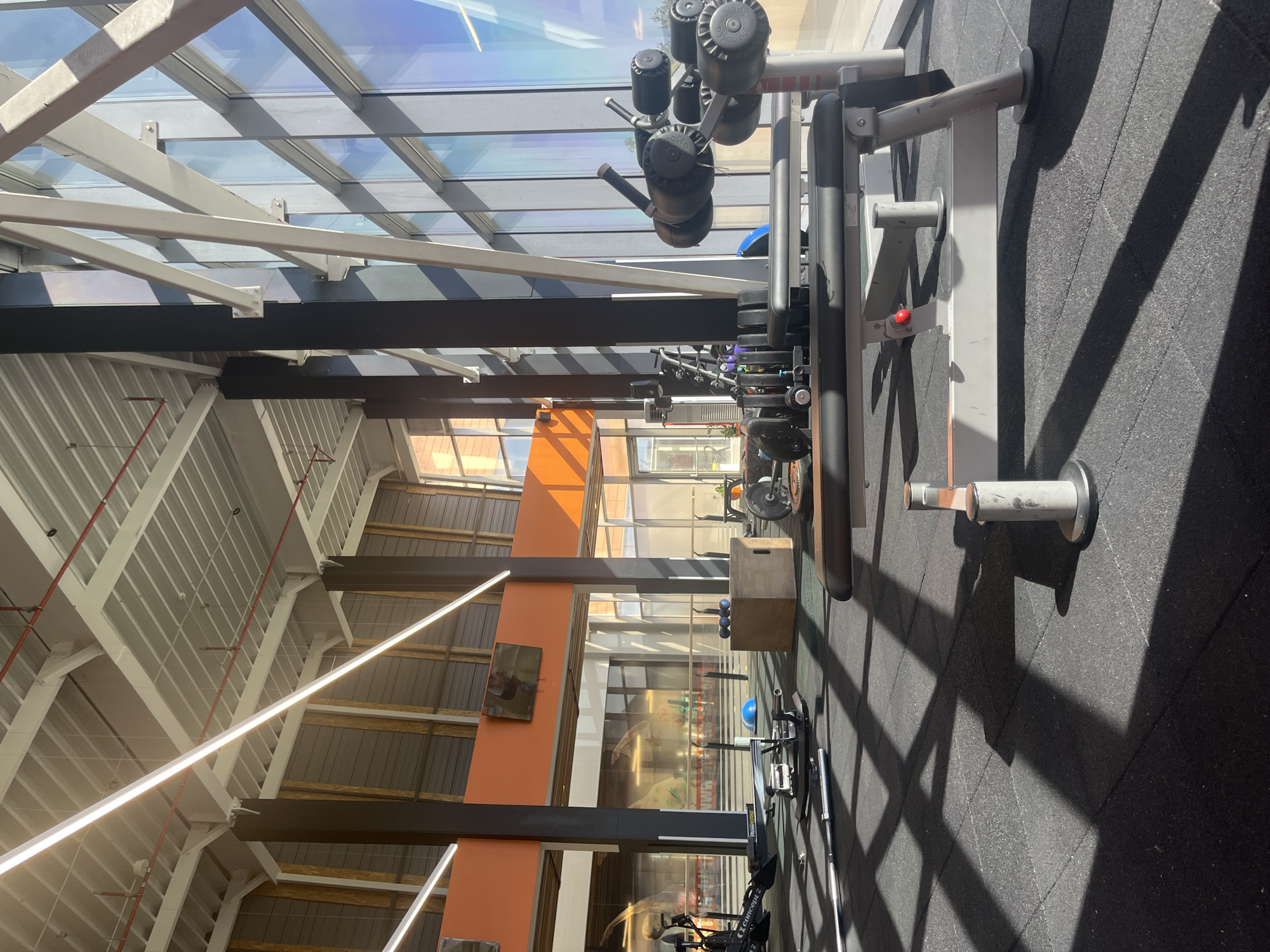 The image shows an interior view of a gym, possibly in Plovdiv, Bulgaria, though the location is not definitively confirmed. The focus is on gym equipment, including weight machines and a plyometric box, placed on a dark rubberized floor. Natural light floods the area through a glass ceiling supported by white and dark steel beams. The walls are a combination of orange and neutral tones, with a TV screen visible on one wall. The gym appears unoccupied at the time the photo was taken.