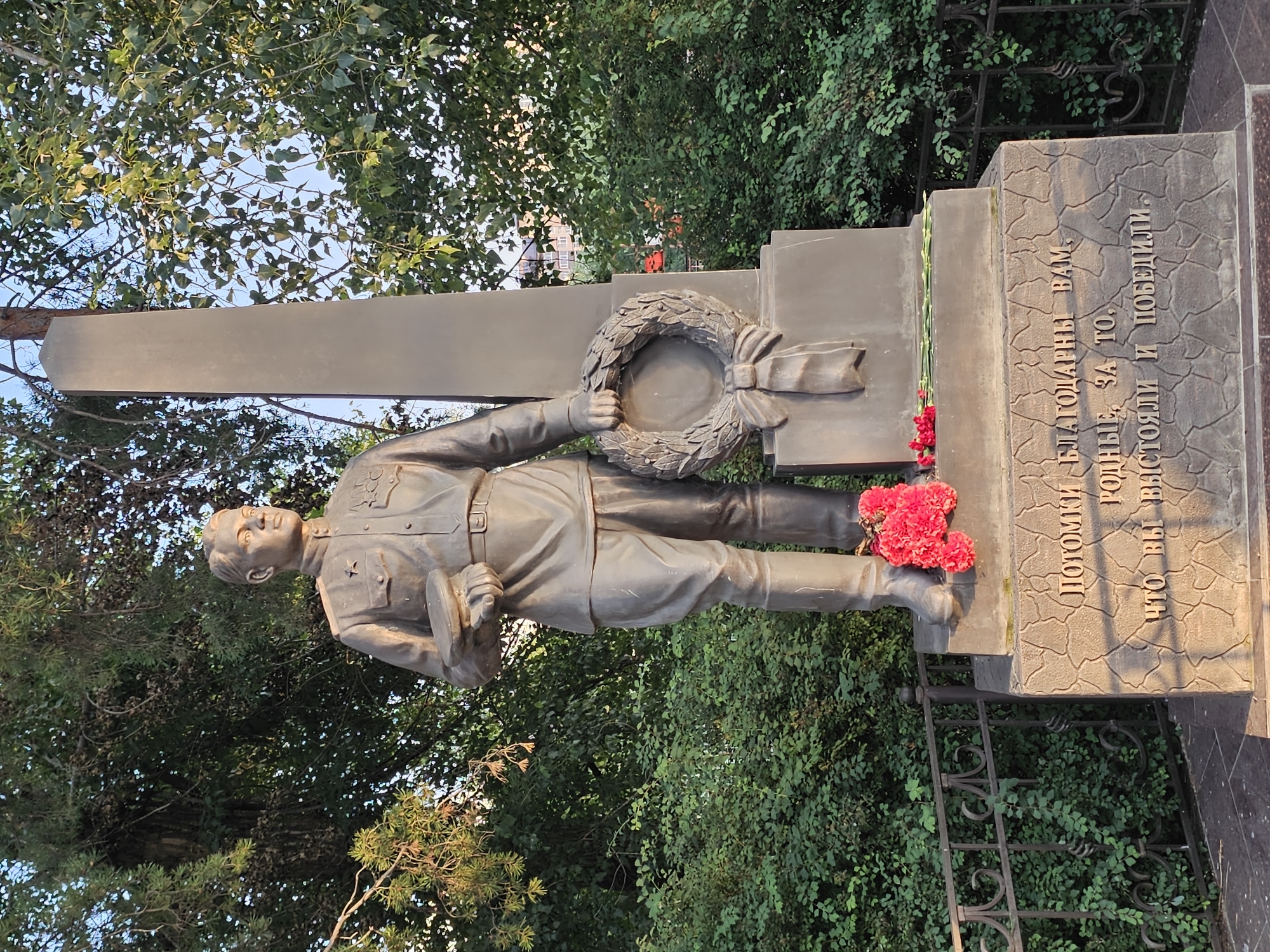 The image shows a bronze monument of a standing Soviet-era soldier holding a cap in his left hand and a laurel wreath in his right. The soldier is wearing a uniform with medals and boots. The monument is located in a park-like setting with lush green trees and bushes. A black metal fence is visible to the left and right of the monument. Red flowers are placed at the base of the monument, which is inscribed with text in Russian. This monument is located in the city of Cherkessk, Russia, and is dedicated to the heroes of the Great Patriotic War.
