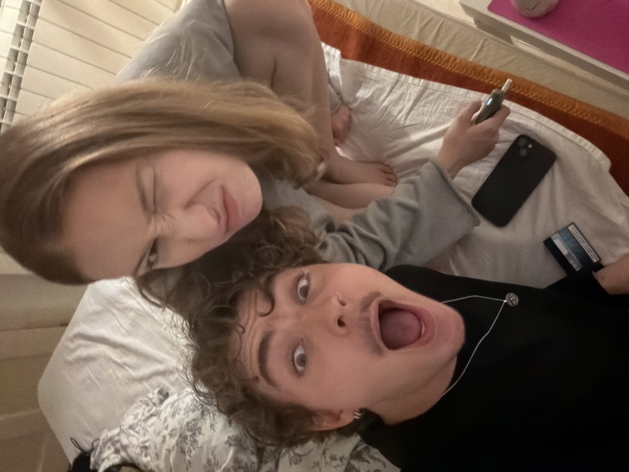 Two individuals are on a bed in what appears to be a bedroom. The person in the foreground, wearing a black long-sleeved shirt and a silver necklace, has curly brown hair and a surprised expression with their mouth open. They are looking directly at the camera. To their right and slightly behind them, another person with long blonde hair is winking and making a humorous face. They are wearing a grey long-sleeved shirt. On the bed, near the person in the foreground, is a black smartphone and a credit card. The person with blonde hair is holding a vape device. The background shows a white radiator. The overall scene suggests a candid, informal moment captured in a residential setting, likely a bedroom. There is no information to suggest the location is in Vracar Urban Municipality, Serbia.