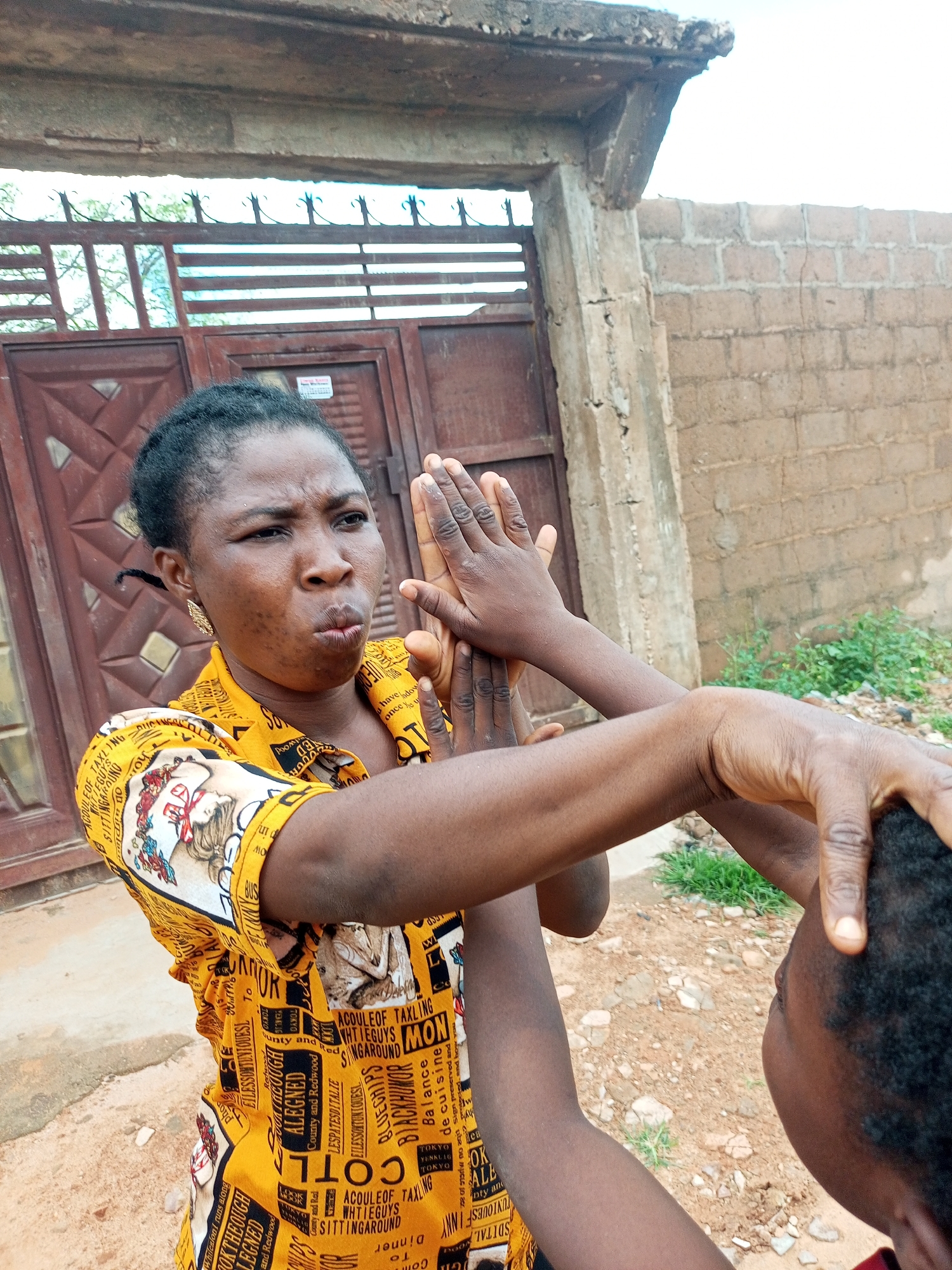 A woman wearing a yellow and white patterned shirt stands outdoors in what appears to be a courtyard or alleyway. She has dark skin and her hair is pulled back. Her face is turned to her right, with her mouth slightly open and her eyebrows furrowed, suggesting she is speaking or reacting to something. Her hands are raised in front of her face, with her palms facing each other. Another person's hands are visible in the foreground, reaching towards the woman's head.
The background includes a weathered, brown metal gate with decorative elements, a concrete structure, and a brick wall. Some green vegetation is visible on the right side of the frame. The ground is covered in dirt and small stones. The overall impression is that of an interaction taking place in a simple, outdoor urban environment. The presence of the gate and the building structures suggest a residential or communal area. The lighting indicates it is daytime.