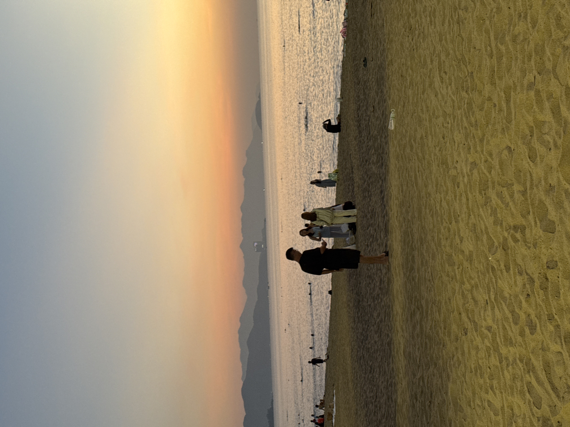 The image captures a serene beach scene in Nha Trang, Vietnam, during the golden hour. In the foreground, rippled sand stretches out, showing footprints and a discarded plastic bottle. A man in a black t-shirt and shorts stands facing away from the camera, while a small group of people, including two women in light-colored clothing, are engaged in conversation to his right.
Further back, the gentle waves of the ocean meet the shore. Several individuals are scattered across the beach and in the water, some swimming, others relaxing. The sea extends to the horizon, where a range of hazy, layered mountains is visible against a gradient sky transitioning from a soft orange near the horizon to a pale blue overhead. The atmosphere is peaceful, with the warm light of sunset bathing the entire scene.