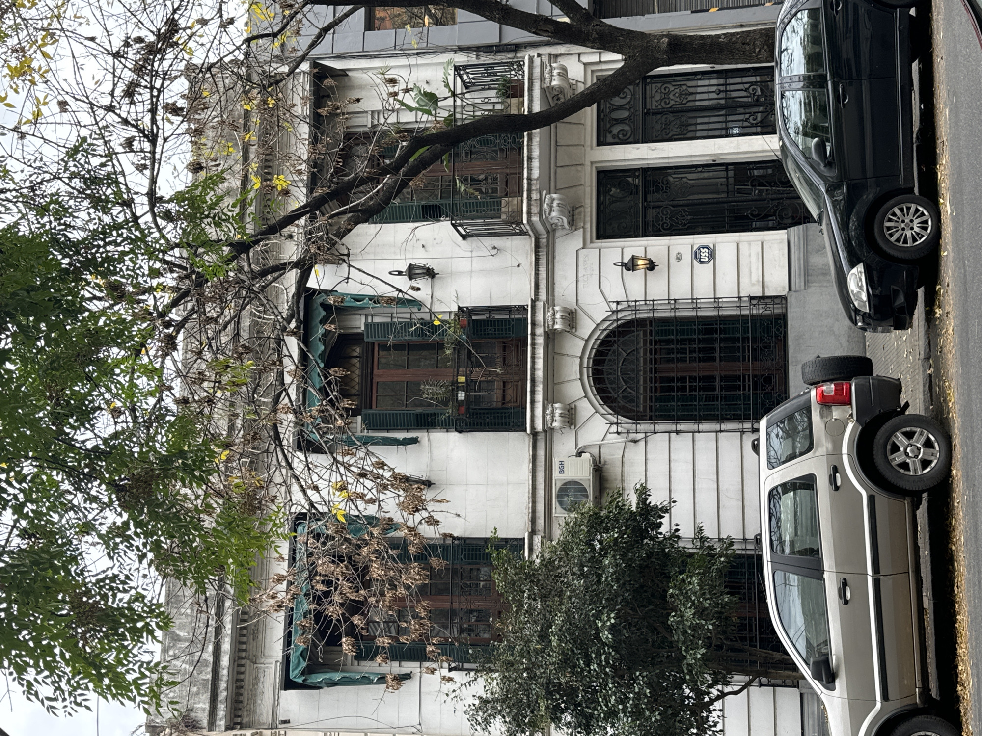 A street view in Buenos Aires, Argentina, showcases a three-story white building with detailed architectural features, including arched windows, ornate molding, and green awnings over some windows. The facade suggests an older, possibly historic, building. A dark car is parked directly in front of the building on the right side of the image, partially obscuring the entrance. A silver SUV with a spare tire mounted on the rear is parked further down the street. Trees with both green and bare branches partially obscure the view of the building.