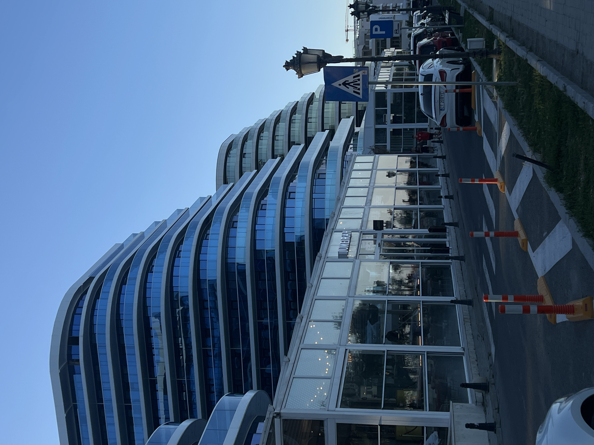 The image depicts a modern, multi-story building with a distinct curved architectural design featuring balconies with blue glass railings. In front of this building, at street level, is a commercial space with large glass windows, possibly a restaurant or shops, as indicated by the text "JUMEIRAH" visible on one of the glass panes. The street in front is paved, with a pedestrian crossing marked by white stripes and orange and white traffic cones. Several cars are parked along the right side of the street, and a street lamp with an ornate design stands next to a pedestrian crossing sign. The sky is clear and blue, suggesting it is daytime, likely late afternoon given the lighting. The scene appears to be a well-maintained urban environment, possibly along a waterfront or promenade, given the sleek, contemporary design of the building and the presence of outdoor seating areas implied by the glass-fronted establishments. The building's design is reminiscent of waves or cascading structures. In the background, construction cranes are visible, indicating ongoing development in the area. The overall impression is one of a modern, possibly resort-style, location in Năvodari, Romania.