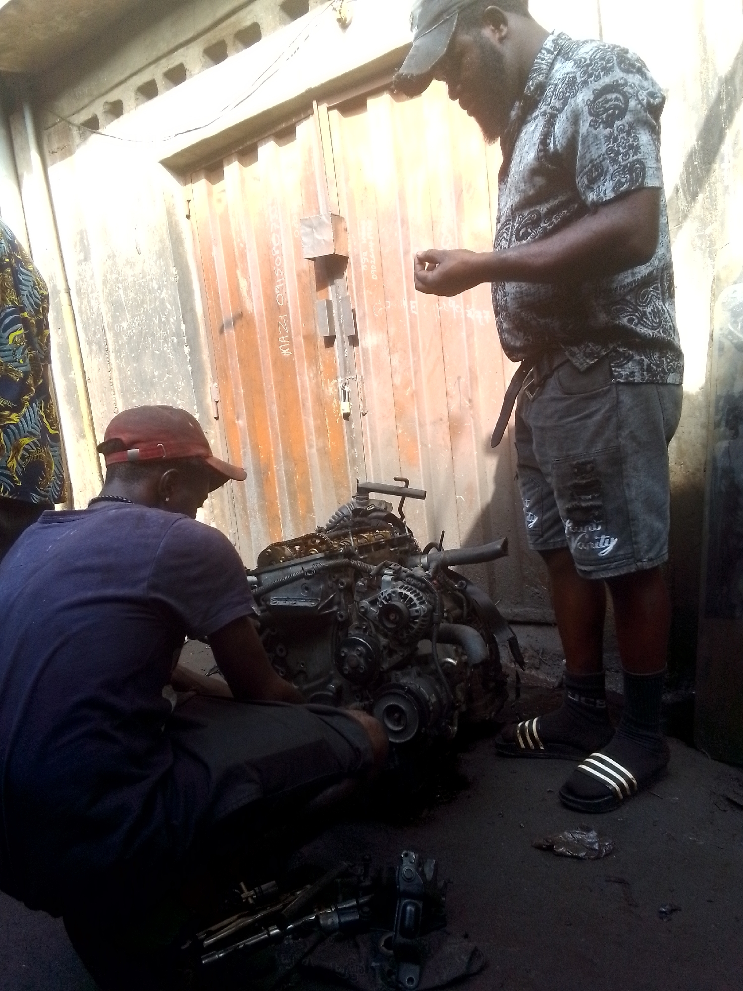 Two men are working on a car engine in what appears to be an outdoor auto repair shop. The man on the left is kneeling and focusing on the engine. The man on the right is standing and looking down at the engine. Tools and parts are scattered on the ground around the engine. The backdrop is an industrial setting, possibly in Onitsha, Nigeria, suggested by the typical environment for such repair businesses.