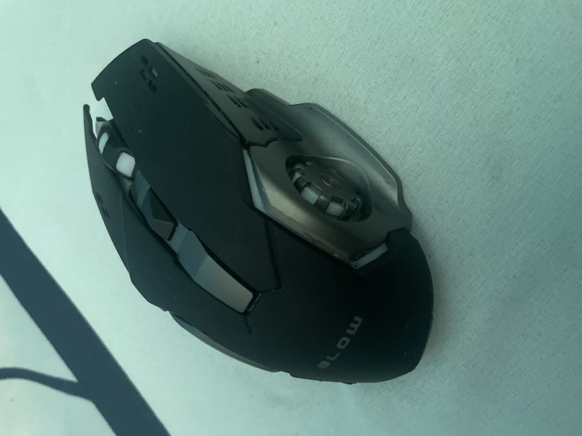 A black gaming mouse with silver accents and the word "BLOW" embossed on its side is placed on a light blue, textured surface. The mouse has a futuristic design with angular lines and visible buttons and a scroll wheel. The surface appears to be a cloth or fabric. The lighting suggests it is indoors, with a shadow cast from an unseen object in the upper left corner. There is no indication of the location being Kołobrzeg, Poland, as no landmarks or identifying features are present in the image.