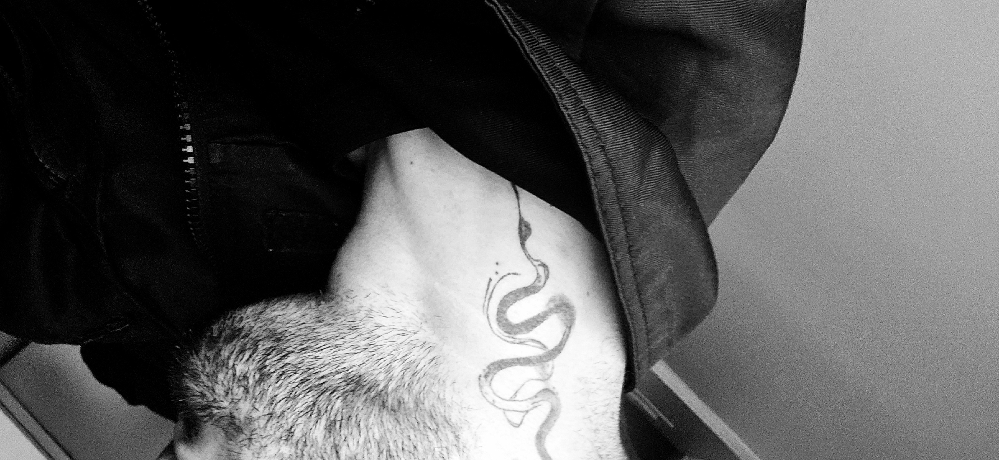 This monochrome, close-up photograph is rotated 90 degrees counter-clockwise, showing the side profile of a person's neck and upper back. The subject, likely male, has short, stubbly hair on the back of their head and neck. A prominent snake tattoo, rendered in an outline style, is visible on the side of their neck, accompanied by smaller dot details. The person is wearing a dark, possibly black, jacket or garment, with a discernible zipper and textured fabric visible on the shoulder area. The background is a plain, light-colored wall or surface, suggesting an indoor setting. No specific activity or event is taking place; the image is a static portrait, emphasizing the tattoo and the texture of the hair and garment. The lighting is soft, creating subtle contrasts in the black and white palette. No emotions are discernible, and no text is visible. This image was captured in Plovdiv, Bulgaria.