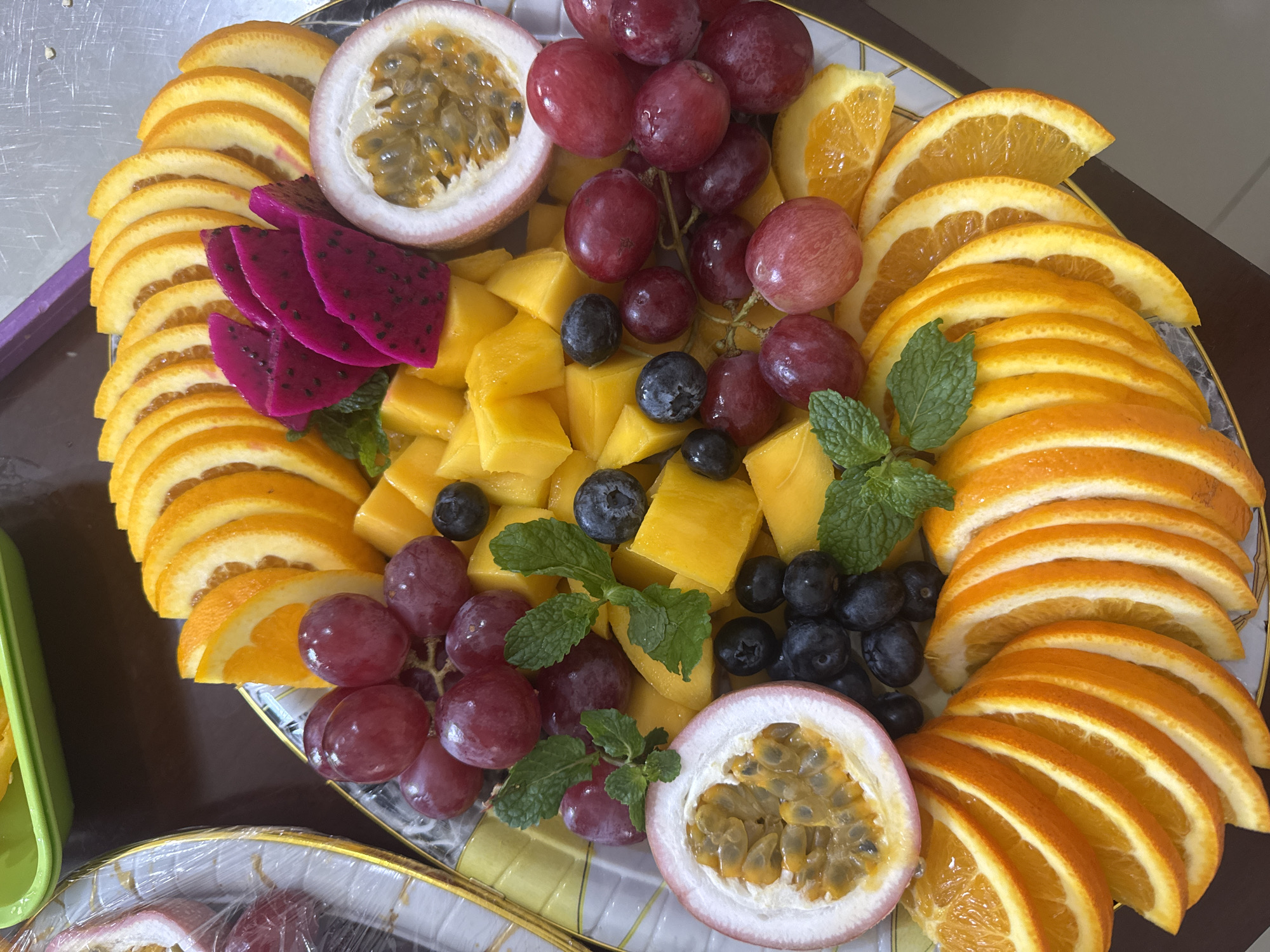 A platter of fruit is centrally composed. The fruit variety includes segments of orange, cubes of mango, red grapes, blueberries, passion fruit cut in half, slices of dragon fruit, and mint leaves for garnish. The fruit appears arranged aesthetically on a white plate with a gold trim. The location is not evident from the image, and there is no indication that the image was taken in Kuta Selatan, Indonesia. There are no people shown in the image.