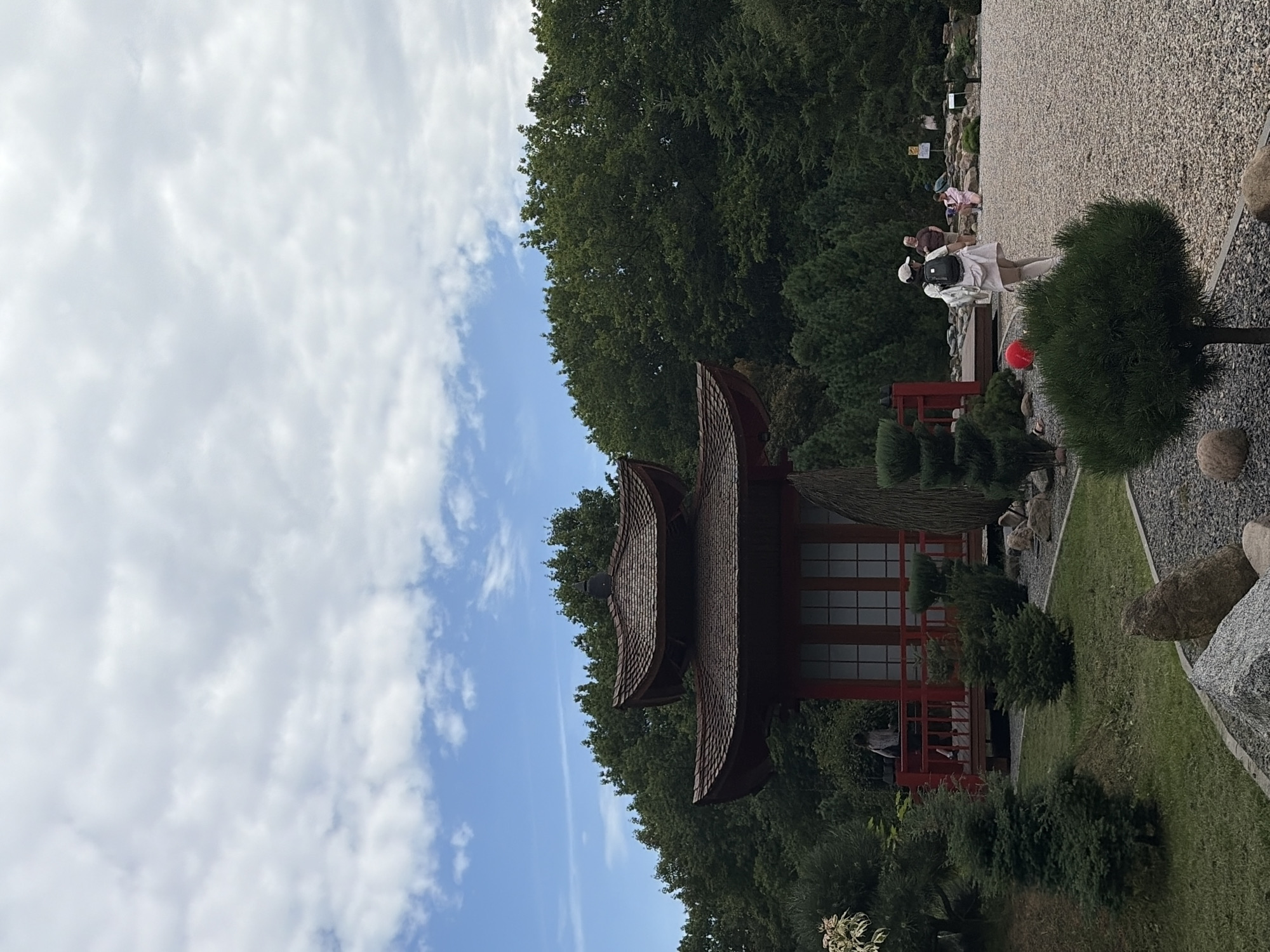 In an outdoor setting with a cloudy sky, a Japanese-style building with a red exterior and tiered, dark brown roof is central to the scene. The building has white-paned windows and a red railing on a raised porch. In front of the building, a person wearing a white outfit and a backpack walks away from the camera on a gravel path. To the left of the building, there is a grassy area with manicured bushes and trees, including a small evergreen in the foreground. Further in the background, more trees form a dense green wall, and several other people are visible, some standing near rocks and one kneeling.