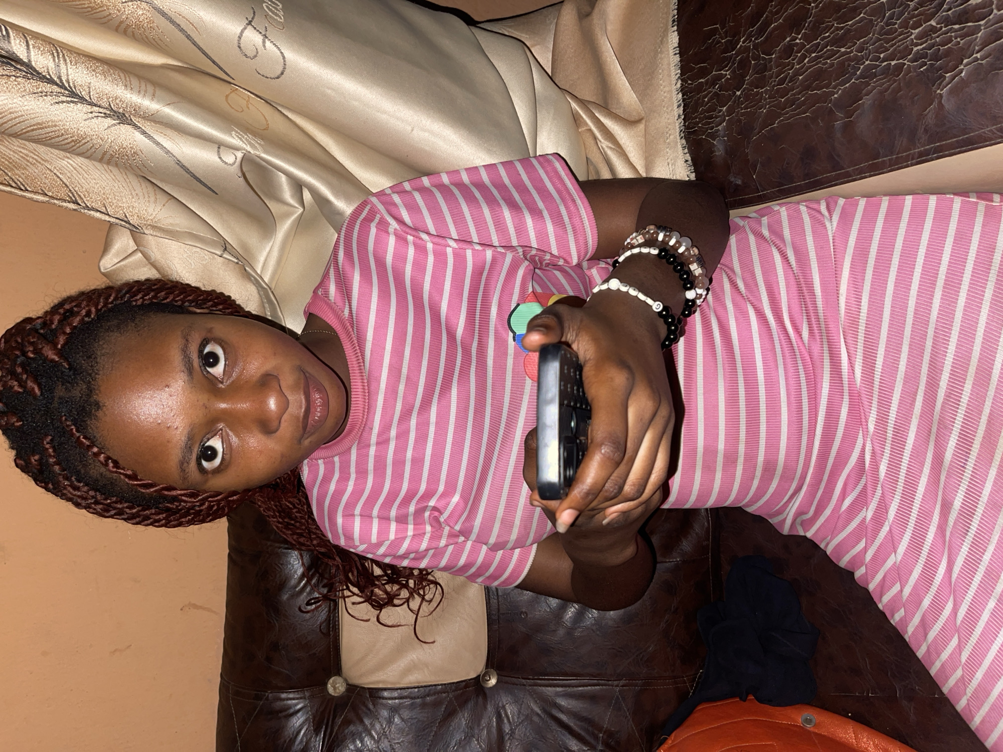 A woman lies on a brown leather couch with a tan patterned pillow behind her. She wears a pink and white striped dress and several bracelets on her left wrist. She holds a black remote control in her left hand, aimed towards the viewer. She has dark hair braided with brown highlights and a direct gaze. A black piece of fabric and an orange object are visible on the couch near her lower body. The location appears to be indoors. There is no information in the image to indicate that this is Muchiya, Nigeria.