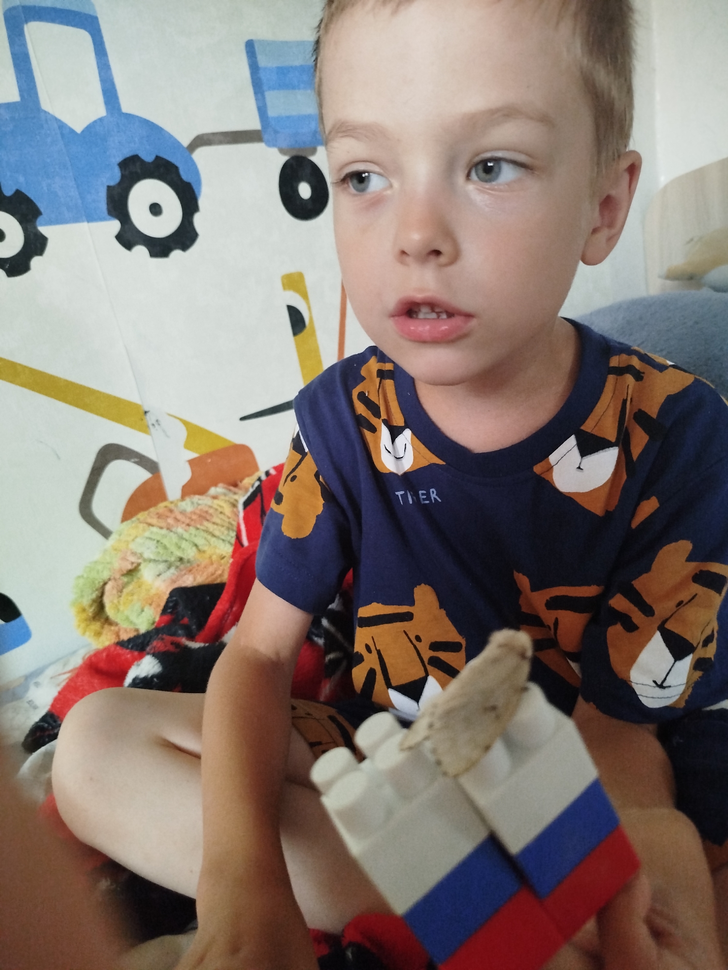 The image shows a close-up of a young boy sitting cross-legged indoors, possibly on a bed, with a blanket partially visible. He is wearing a dark blue t-shirt with an orange tiger print. In his hands, he is holding a construction toy block with red, white, and blue colors, resembling the Russian flag. A small moth is perched on top of the toy block. The background features a curtain or wall covering with a pattern of blue tractors and yellow stripes. The setting appears to be a child's room or living space. There is no visible indication of the location being specifically in the city of Zavodoukovsk, Russia.