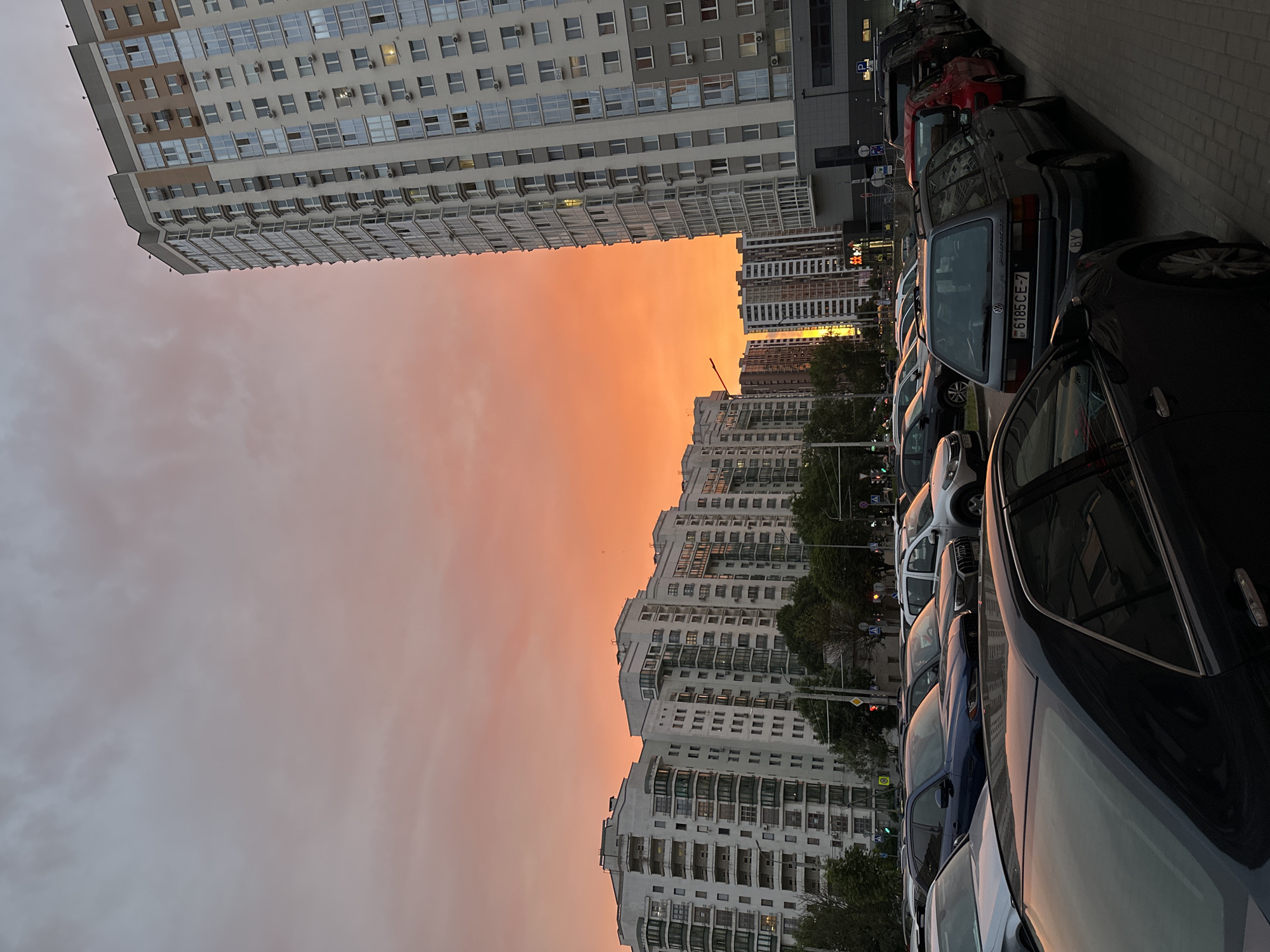 This media file depicts a street scene in Minsk, Belarus, during a vibrant sunset. The sky is a prominent feature, displaying a gradient of fiery orange and soft pink hues, indicative of the late evening light.
The scene is dominated by several large, multi-story residential buildings that line the street. These buildings are modern in design, with numerous windows and balconies. In the foreground, a row of parked cars is visible, facing away from the viewer. The cars vary in color and model, with some of the license plates featuring Cyrillic script, consistent with the location.
One of the cars, a light blue hatchback, has a license plate that appears to read "6185 CE-7" with "BY" below it, which likely indicates Belarus. There are also streetlights and traffic signals present, suggesting this is a developed urban area. Trees are interspersed between the buildings and along the street.
The overall atmosphere of the image is calm and serene, with the dramatic sunset providing a striking backdrop to the urban landscape. The activity taking place is the natural transition from day to night, with the setting sun casting long shadows and bathing the scene in warm, colorful light. No people are clearly visible, and the focus is on the architecture, the sky, and the parked vehicles. The time of day is clearly sunset or dusk, and the weather appears clear, with the colorful sky suggesting good visibility.