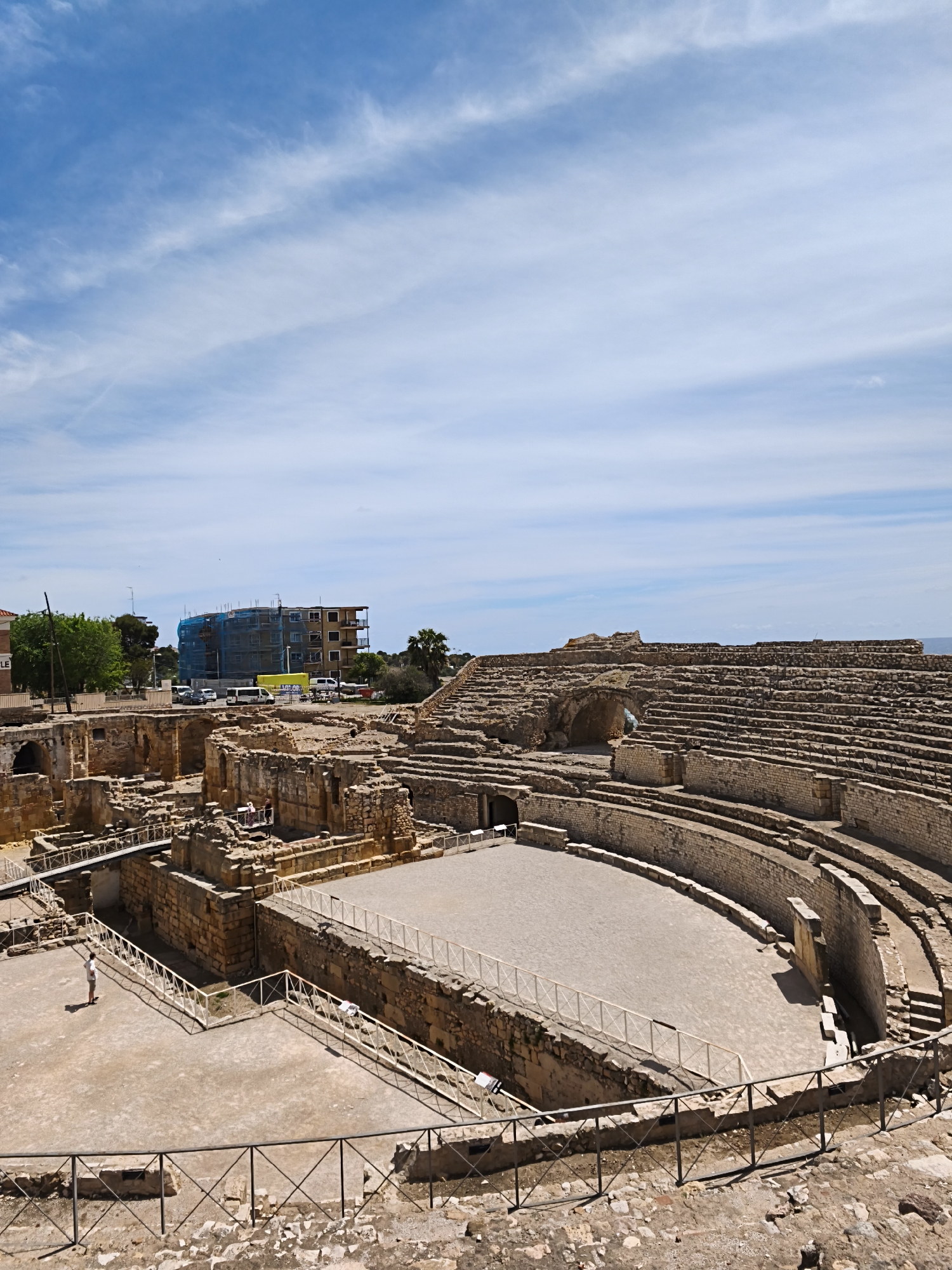 The image shows the ruins of the Roman amphitheater in Tarragona, Spain, under a bright blue sky with scattered clouds. The amphitheater’s stone structure is well-preserved, revealing tiers of seating overlooking a flat, sandy arena. A low fence surrounds the arena. A solitary person stands near the lower left perimeter of the arena. In the background, beyond the amphitheater's perimeter, are modern buildings and a street with parked cars. A building is covered in blue scaffolding.