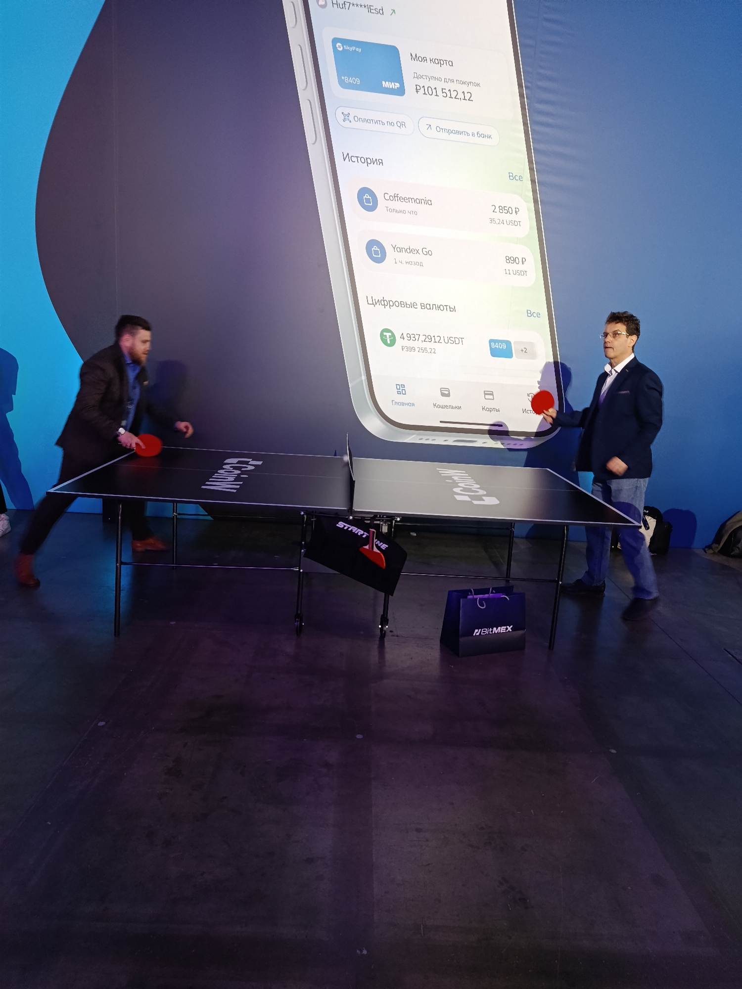 Here is a description of the image:
The image captures a scene where two men are engaged in a game of table tennis. One man is wearing a dark suit and brown shoes. He is in motion with his arm extended, ready to hit the ball. The other man is wearing a blue suit and jeans. He is holding a red paddle. A black table tennis table with white logo on it sits between them.
Behind the table, a large display is showing the interface of what appears to be a mobile banking application. The application shows various details like account balance, recent transactions (like "Coffeemania" and "Yandex Go"), and digital currency balances.
A black bag with the logo "BitMEX" is on the floor. A black and red object with "START NE" printed on it is also visible.
Given the Russian text in the application interface, the image likely originates from Russia. The text "Huf7****IEsd" and some Russian text on the display suggest a connection to digital finance. There is no direct indication this is in Khimki, Russia, but it cannot be ruled out based on the image.
