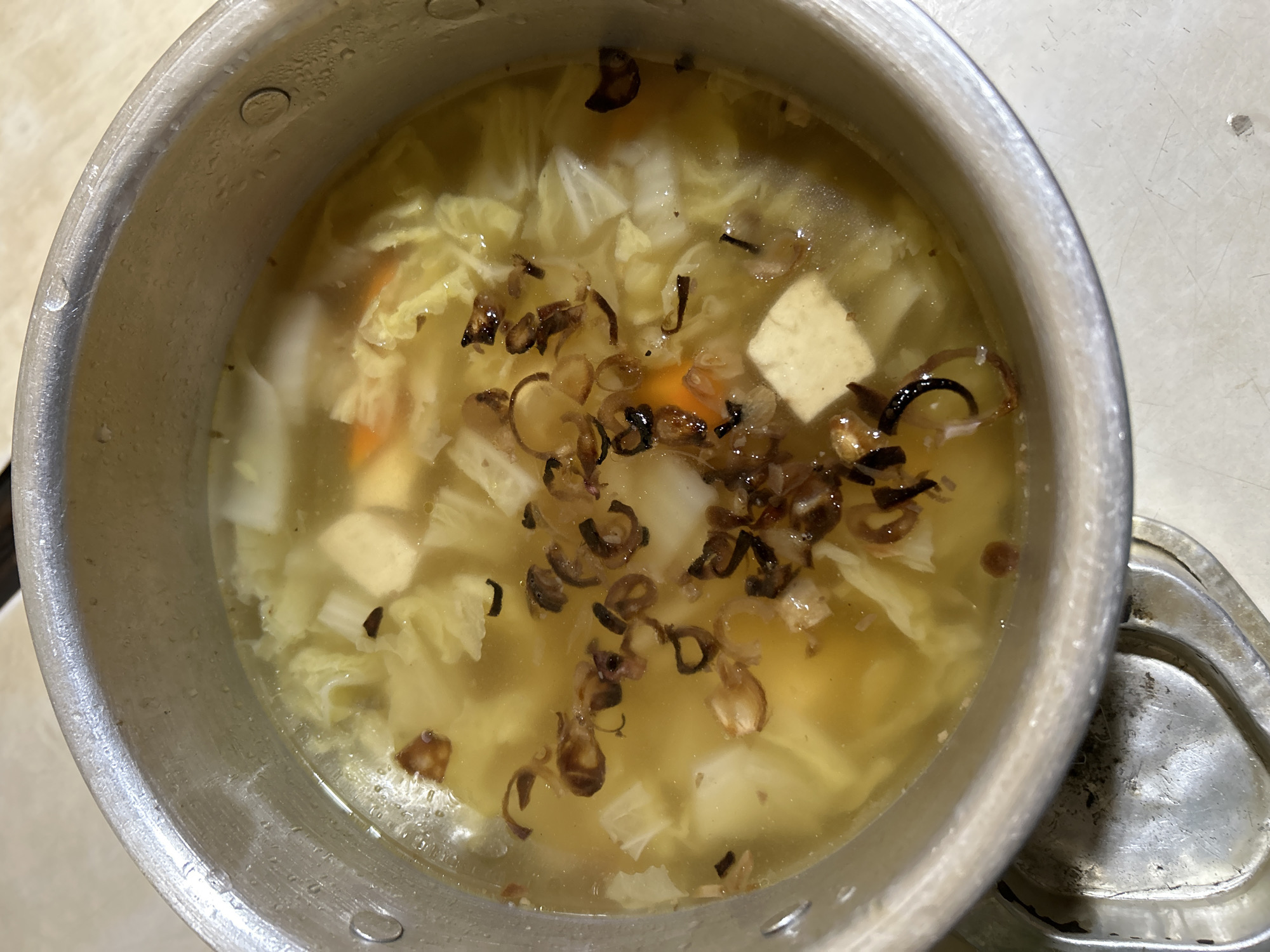 This is a close-up, overhead shot of a pot of clear soup with various ingredients, taken in Depok, Indonesia. The soup contains chunks of white tofu, shredded leafy green vegetables, sliced carrots, and what appears to be scrambled egg. The surface of the soup is garnished with crispy fried shallots. The soup is in a silver metal pot with visible water droplets on the inside rim. The pot is placed on a metallic surface, possibly a stove or a kitchen counter. The overall lighting suggests an indoor setting, likely a kitchen environment.