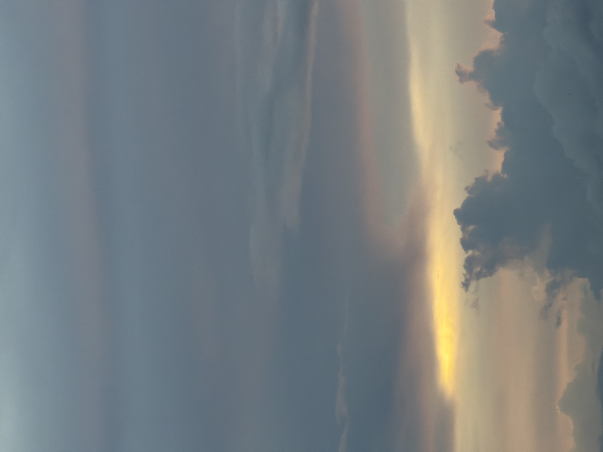 The image is a photograph of a sky with clouds, taken at an angle looking upwards. The sky is a gradient of blue and grey, with streaks of orange and yellow light appearing near the horizon, suggesting either sunrise or sunset. The clouds are varied in density and shape, with some appearing wispy and others dense and dark. The overall impression is a dramatic sky scene, characteristic of twilight or a storm approaching. The photograph does not contain any identifiable landmarks or structures that would specifically place it within Na Thung, Thailand, beyond it being a sky photograph.