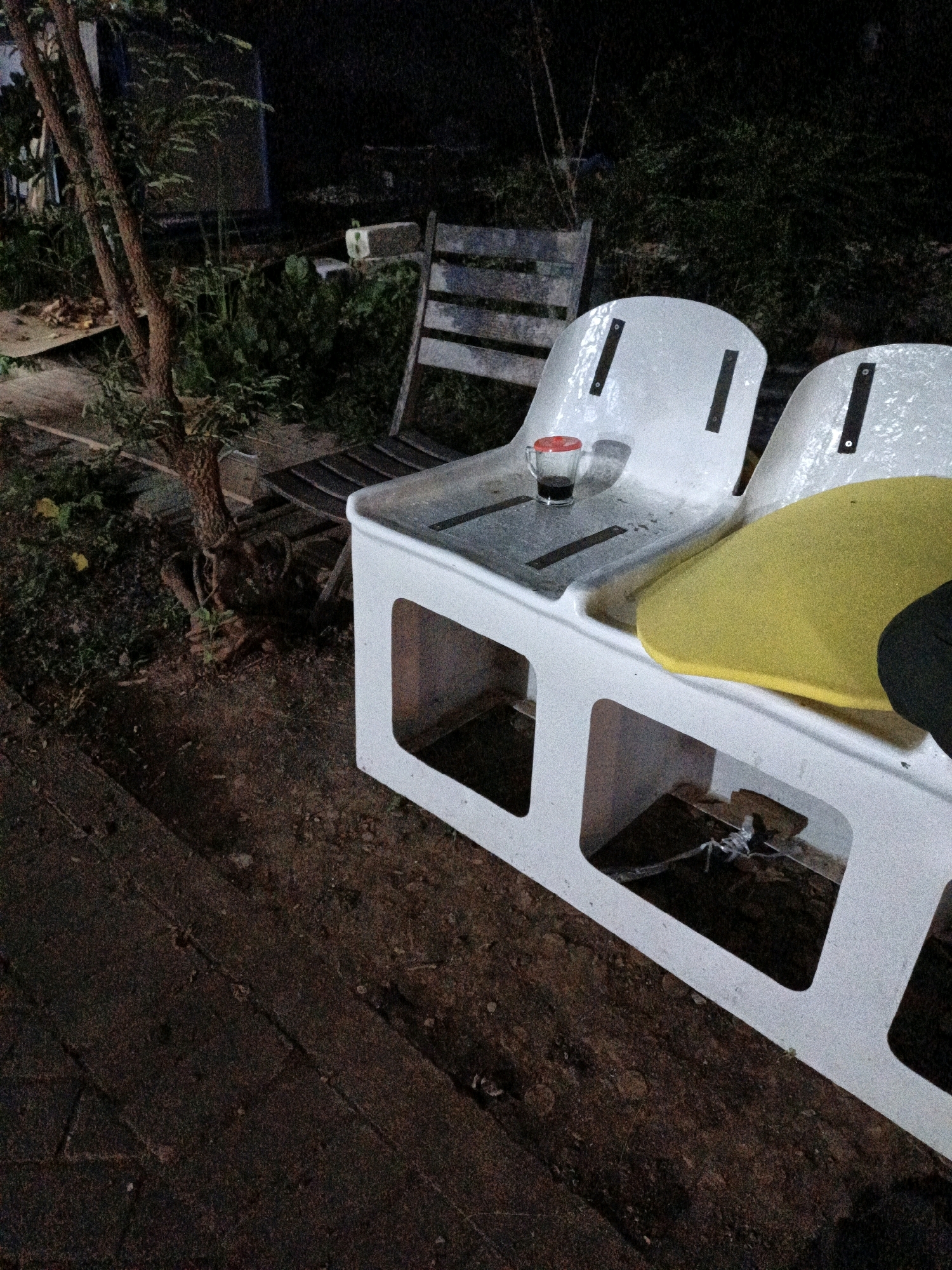 The image captures a nighttime scene in Cikal, Indonesia, featuring outdoor seating in a somewhat disheveled environment. In the foreground, a white, rectangular bench with two seats is prominent. One seat is empty, while the other has a bright yellow cushion and a black object, possibly a bag or garment, partially visible. On the left seat, a small glass cup containing a dark liquid and topped with a red lid sits on the surface.
Behind the bench, a weathered wooden chair with horizontal slats is positioned. To the left of the bench, a young tree with visible roots grows from the uneven, earthy ground. The surrounding area is overgrown with vegetation, with darker, dense foliage in the background suggesting it's a natural or semi-wild setting. The ground appears to be a mix of soil, dirt, and possibly some scattered leaves or debris. The lighting is dim, indicating it's nighttime, with artificial light sources not clearly discernible, but casting shadows that contribute to the overall mood. The presence of the bench and chair suggests an area for public seating or a communal gathering space.