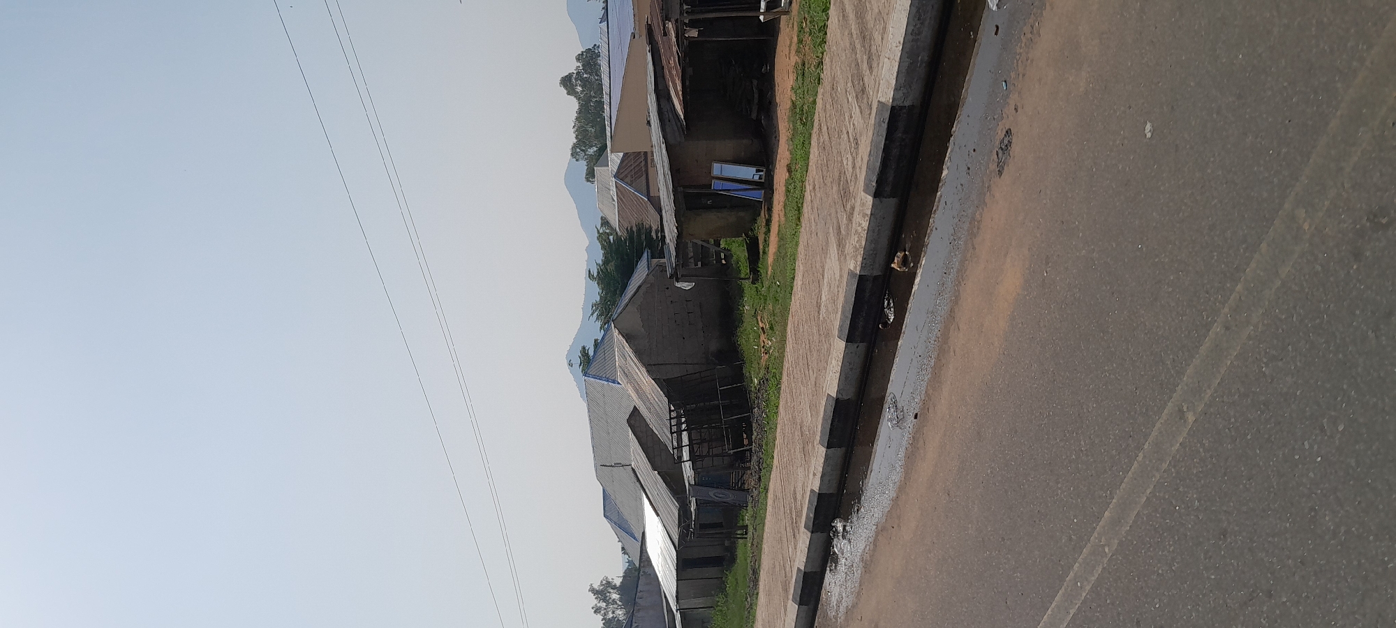 The image shows a roadside scene in Jalingo, Nigeria. Buildings with corrugated metal roofs line a paved walkway, which is separated from a tarmac road by a curb with black and white stripes. Some of the buildings appear to be shops or residences, with windows and open doorways visible. The road shows signs of wear, with some debris and puddles. In the background, lush green vegetation and rolling hills can be seen under a clear, pale blue sky. There are power lines stretching across the sky. The overall impression is one of a rural or semi-urban environment, likely during the daytime due to the bright lighting. No people are visible in the image, and no text is discernible. The scene suggests a typical day in this Nigerian town.