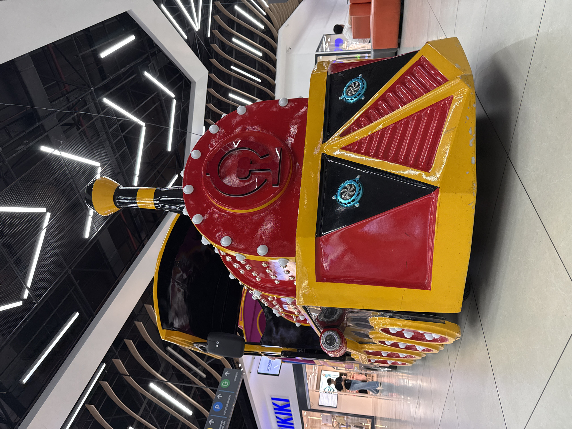 The image shows a colorful, rideable toy train, likely for children, positioned in what appears to be a shopping mall. The train's engine is predominantly red and yellow, with decorative elements and visible wheels. The backdrop features modern mall architecture, with overhead lighting and storefronts. One visible store sign reads "KIKI," and there's a navigation sign indicating "Parking" and other mall directions. People are faintly visible in the background, suggesting a populated indoor environment. Based on the provided text "Tashkent, Uzbekistan," the photo was likely taken in a shopping mall in that city.