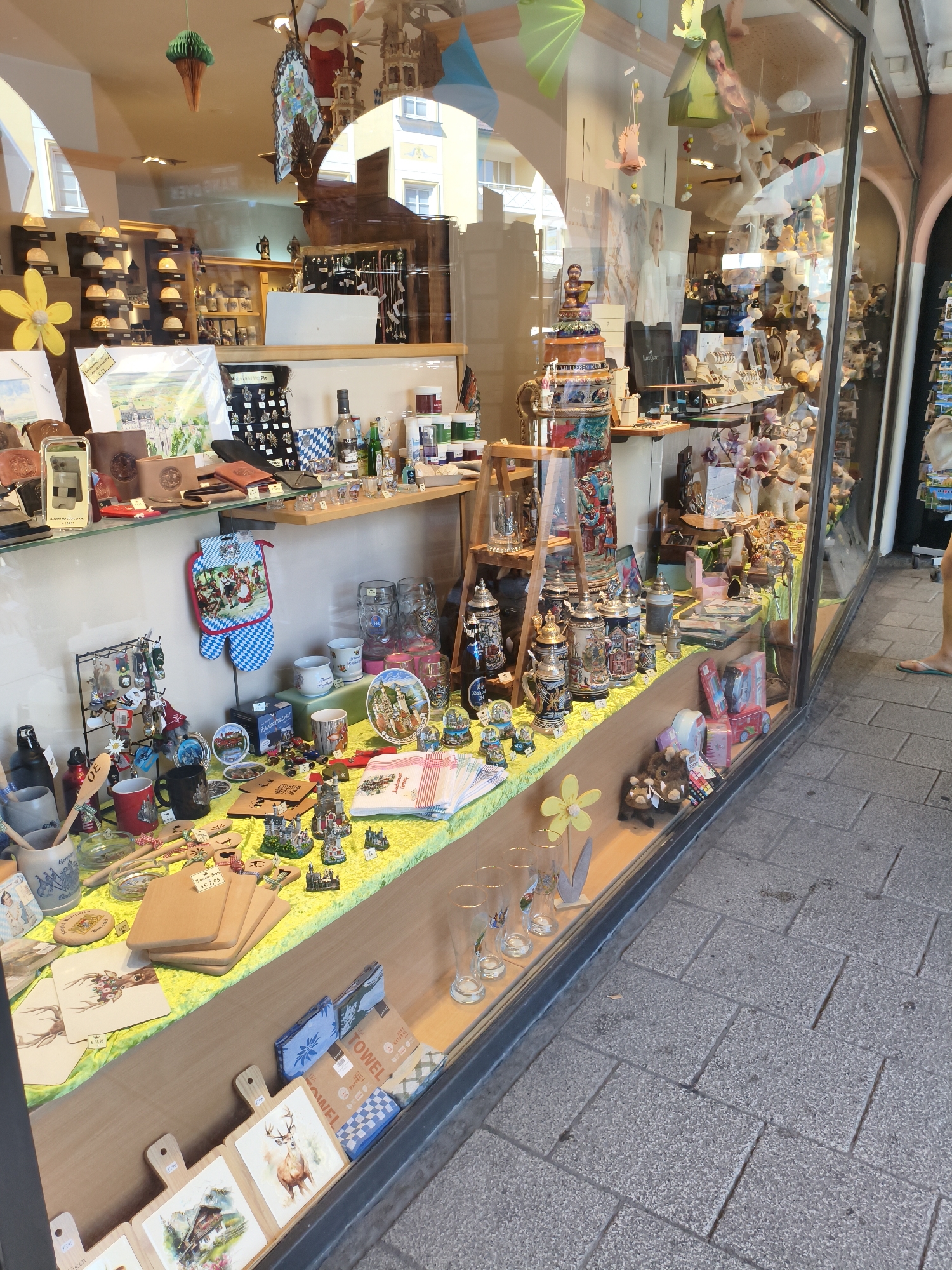 The image shows a street-level display window of a shop in Füssen, Germany. The window is filled with a variety of items, primarily decorative and souvenir-style objects.
On the left side of the display, there are several shelves featuring hats on stands, framed prints of landscapes, and leather goods such as wallets and photo frames. A large yellow artificial flower is prominently displayed. Below this, a table is laden with kitchen items like oven mitts, wooden spoons, cutting boards, and mugs. There are also decorative figurines and small beer steins.
The center of the display showcases a collection of traditional German beer steins of various sizes, along with glass beer mugs. Scattered among these are smaller decorative items, including plates with scenic depictions, and more figurines. A stack of towels is also visible.
The right side of the display features more beer steins, a bottle of liquid, and an assortment of other souvenirs. In the background, through the glass, other parts of the shop interior are visible, including shelves stocked with plush toys and other merchandise, as well as decorative elements like pink magnolias.
The exterior setting shows a paved sidewalk made of grey stone tiles arranged in a herringbone pattern. The shop window is large and made of glass, reflecting the surroundings. A person's legs, wearing a light-colored top and teal flip-flops, are partially visible on the right side of the image, indicating the sidewalk is in front of the shop. The architecture visible through the window suggests buildings with windows and decorative elements typical of a European town.