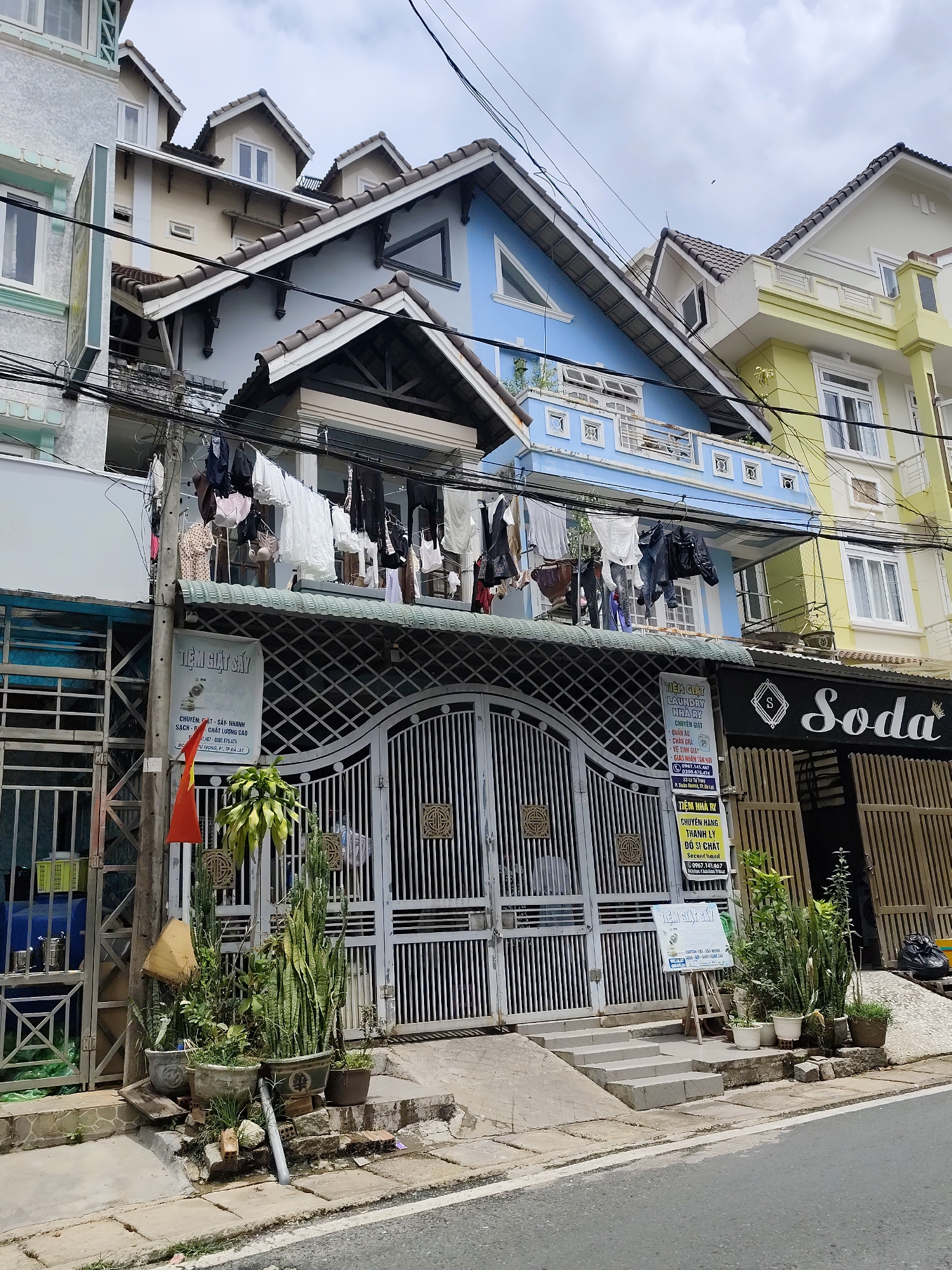 This image captures a vibrant street scene in Da Lat, Vietnam, featuring a row of multi-story buildings under an overcast sky. The central focus is a light blue building with a prominent A-frame roof and a large, ornate metal gate at ground level. Above the gate, clothes are strung on a line, drying in the open air, a common sight in the region.
To the left of the blue building is a lighter, possibly off-white or light green structure, partially visible. To its right, a bright yellow and white building extends further down the street. All buildings feature pitched, tiled roofs, many with dormer windows, lending a distinct architectural style. Numerous utility wires crisscross overhead, adding to the urban landscape.
At street level, in front of the central blue building, several potted plants, including snake plants and cacti, are arranged on the sidewalk. A small red Vietnamese flag with a yellow star is affixed to a utility pole. The sidewalk is made of concrete slabs, leading to a paved road in the foreground.
Several business signs are visible, indicating commercial activity. A large white sign on the left advertises "TIỆM GIẶT SẤY" (Laundry shop) named "NHÀ RY" (House Ry), specializing in fast, clean, high-quality washing and drying, with phone numbers and the address "TỰ TRỌNG, P1, TP ĐÀ LẠT." Another smaller sign for "LAUNDRY NHÀ RY" lists services like washing clothes, bed sheets, shoe cleaning, and home pickup/delivery. Below this, a sign reads "TIỆM NHÀ RY CHUYÊN HÀNG THANH LÝ ĐỒ SI CHẤT Second hand," indicating a second-hand clothing shop. To the far right, a dark sign with white lettering clearly displays the name "Soda" with an 'S' logo.
The scene depicts a typical daytime setting, with no direct sunlight indicating an overcast or cloudy day. No people are visible, but the laundry and business signs suggest daily life and commerce.
